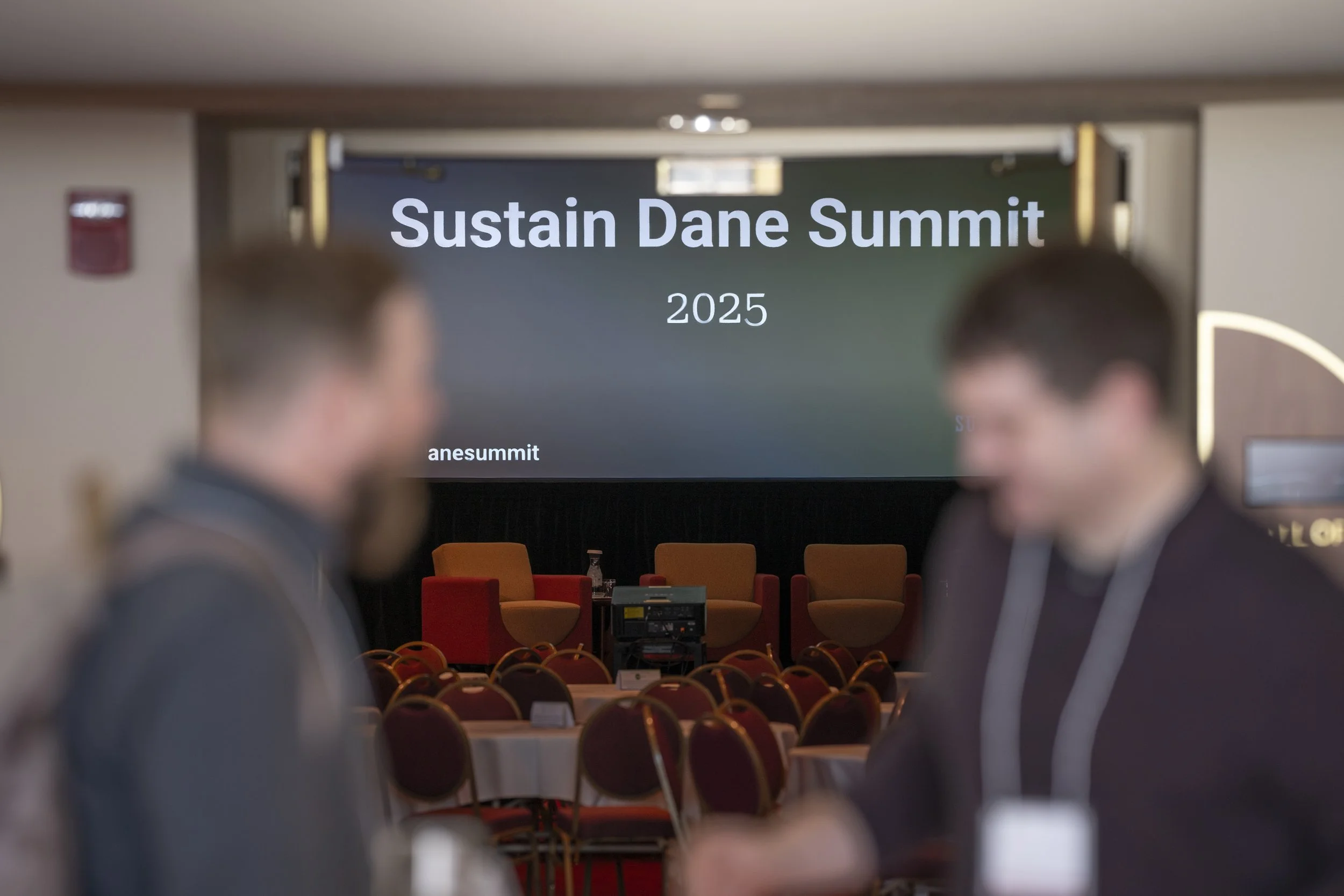 Two people attending the Sustain Dane Summit 2025 conference, with a stage and chairs in the background