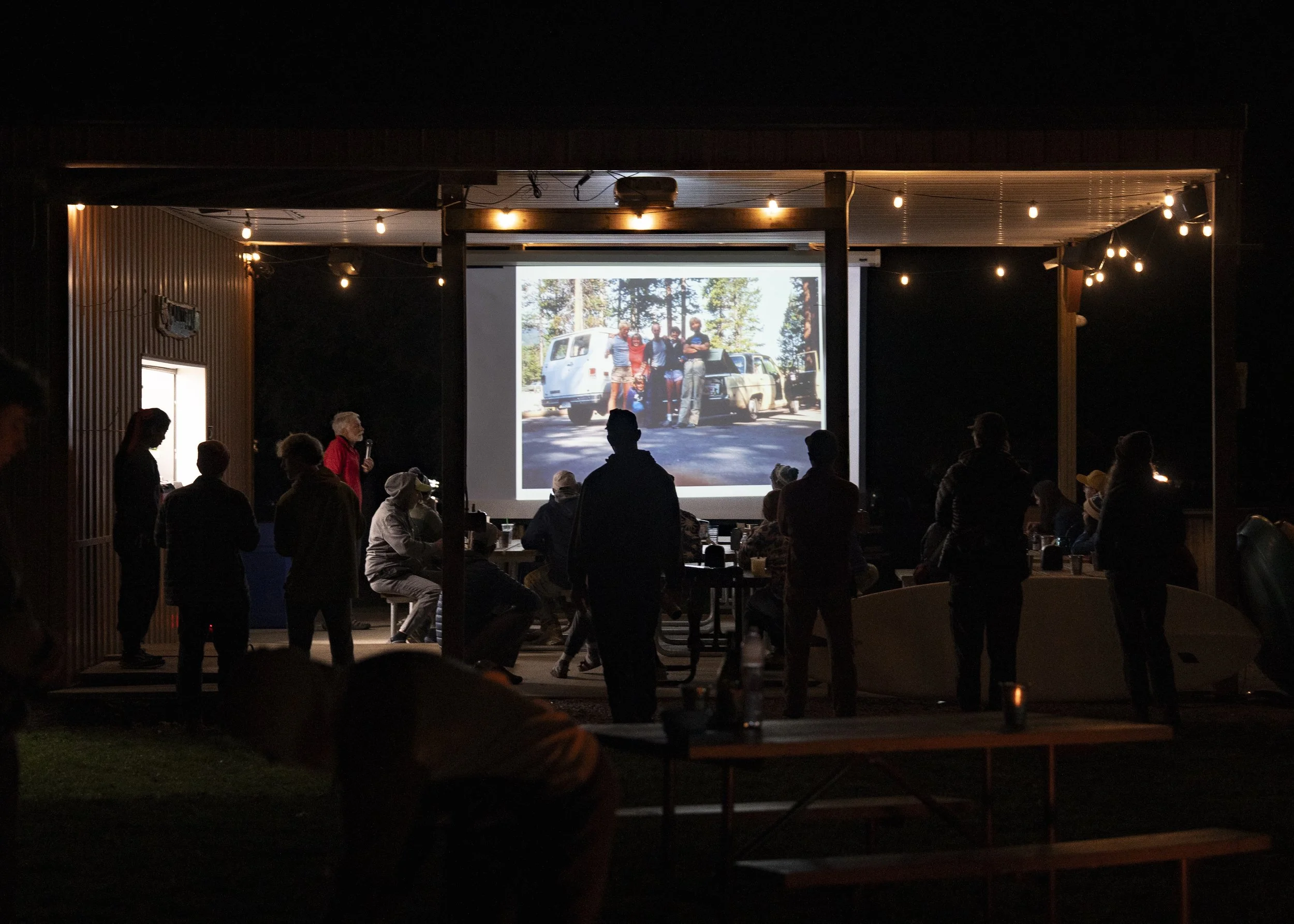 People gathered outdoors watching a movie projection on a large screen at night, with string lights overhead.