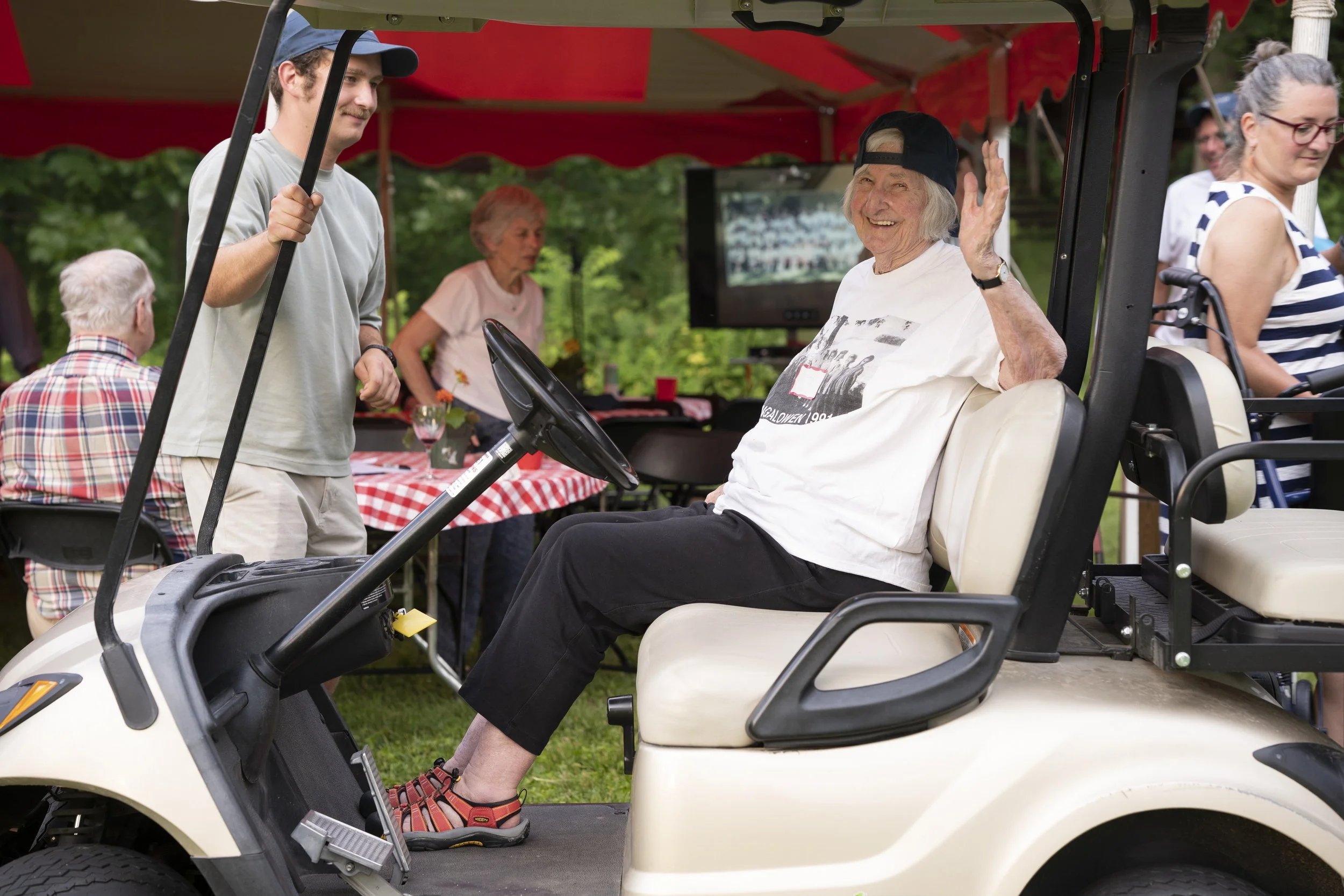 An elderly woman sitting in a golf cart, smiling and waving, at an outdoor gathering with other people and red checkered tablecloths.