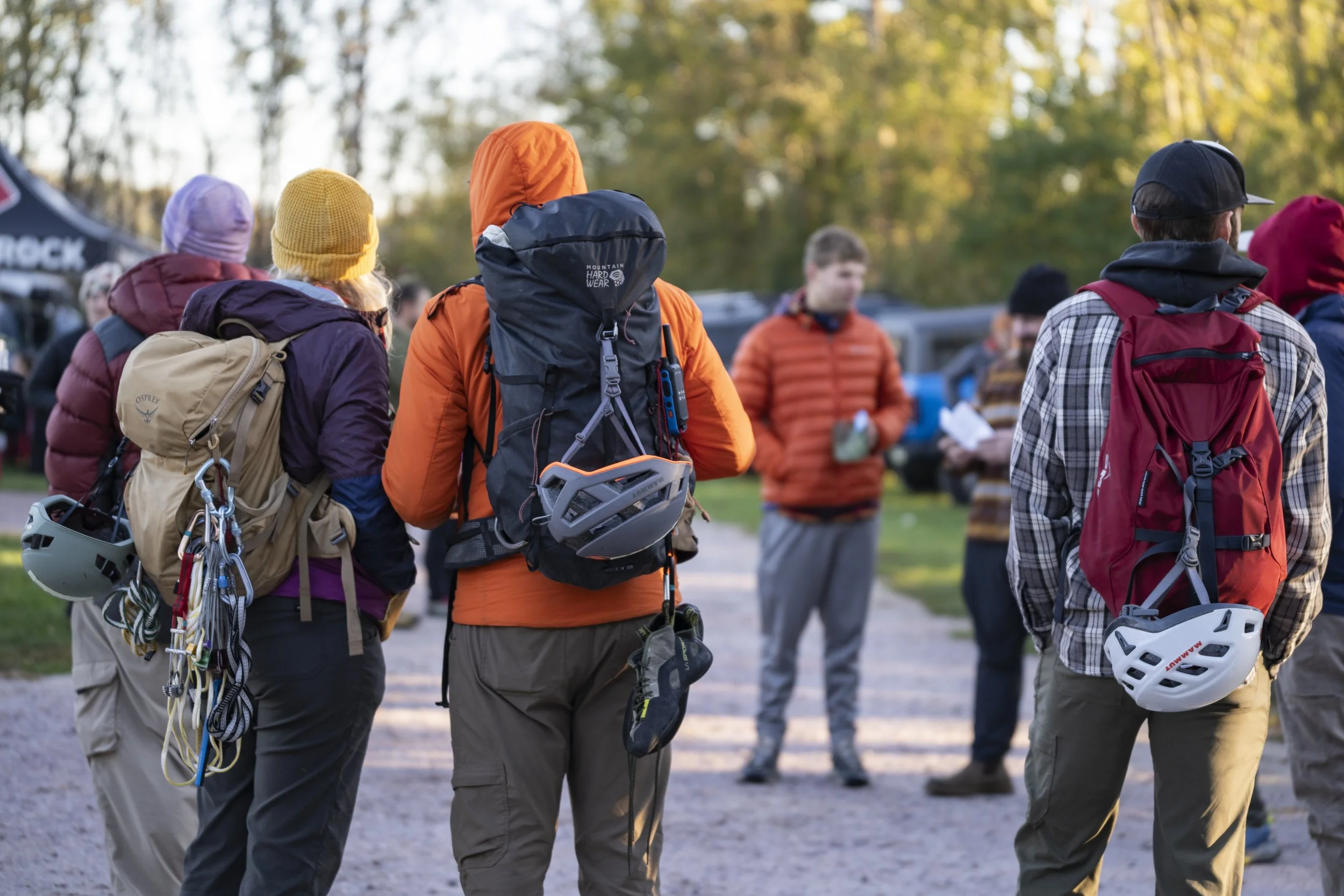 Group of people outdoors, wearing backpacks and warm clothing, likely preparing for a hiking or climbing activity during late afternoon.