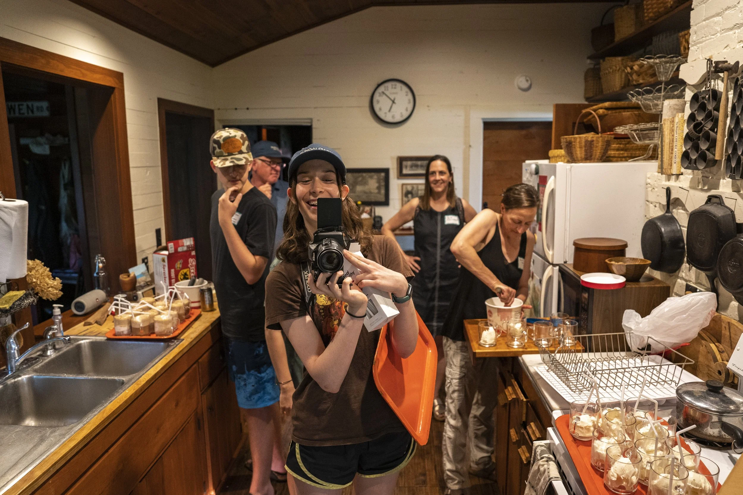 Group of people in a kitchen, with one woman taking a selfie while others prepare food.