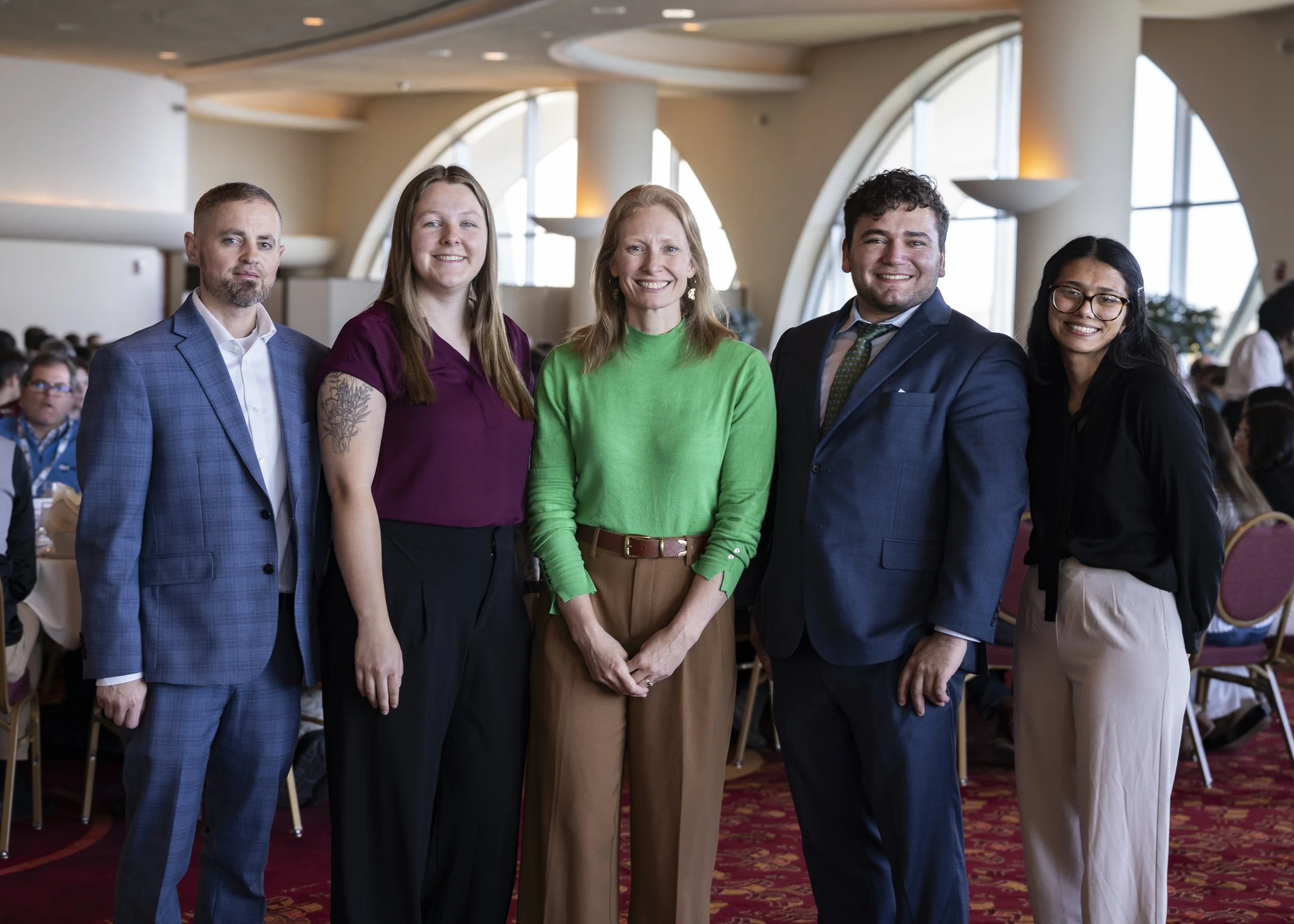 Five individuals standing together indoors, smiling, with a large crowd seated behind them in a banquet hall with high, arched windows.