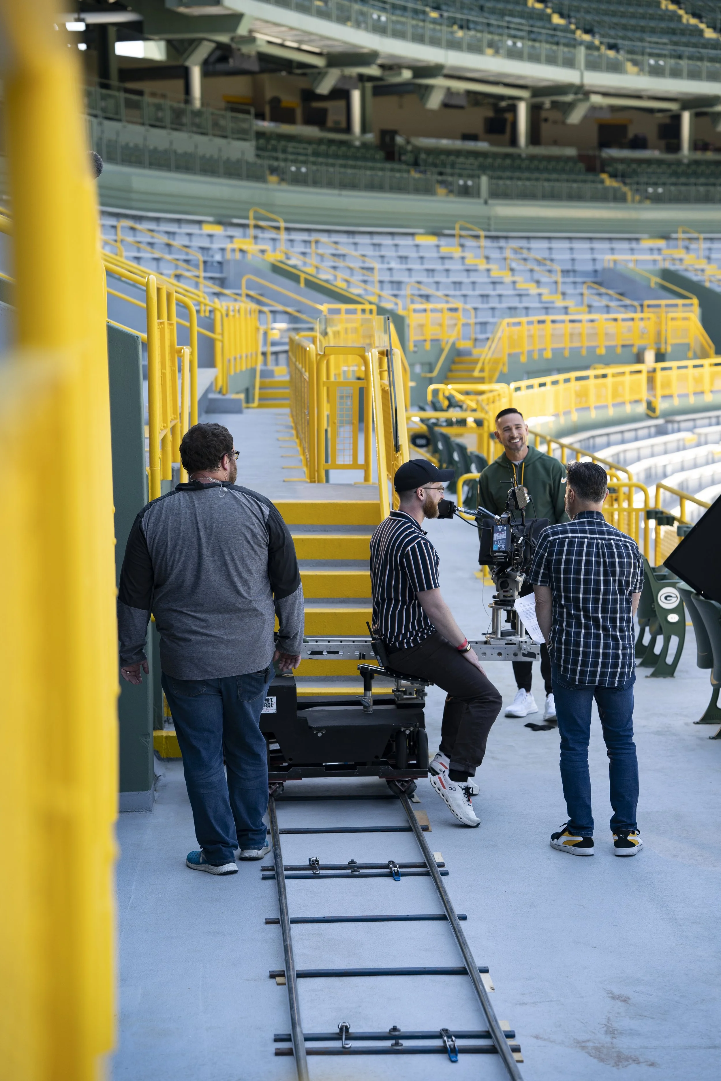 Crew filming at a sports stadium with yellow railings, empty seats, and a track.