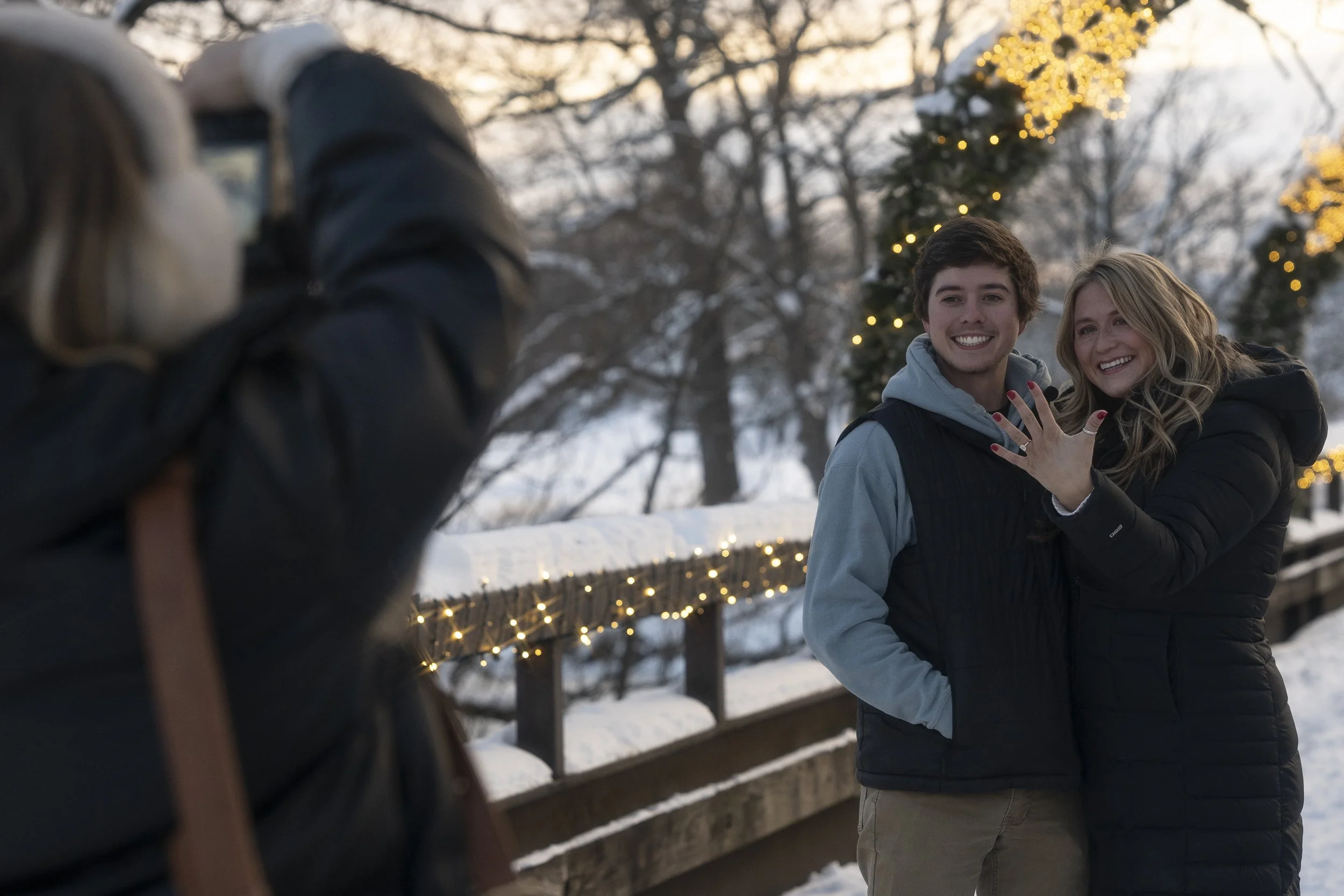 A young man and woman smiling and posing for a photo on a snowy winter day with decorated Christmas lights in the background.