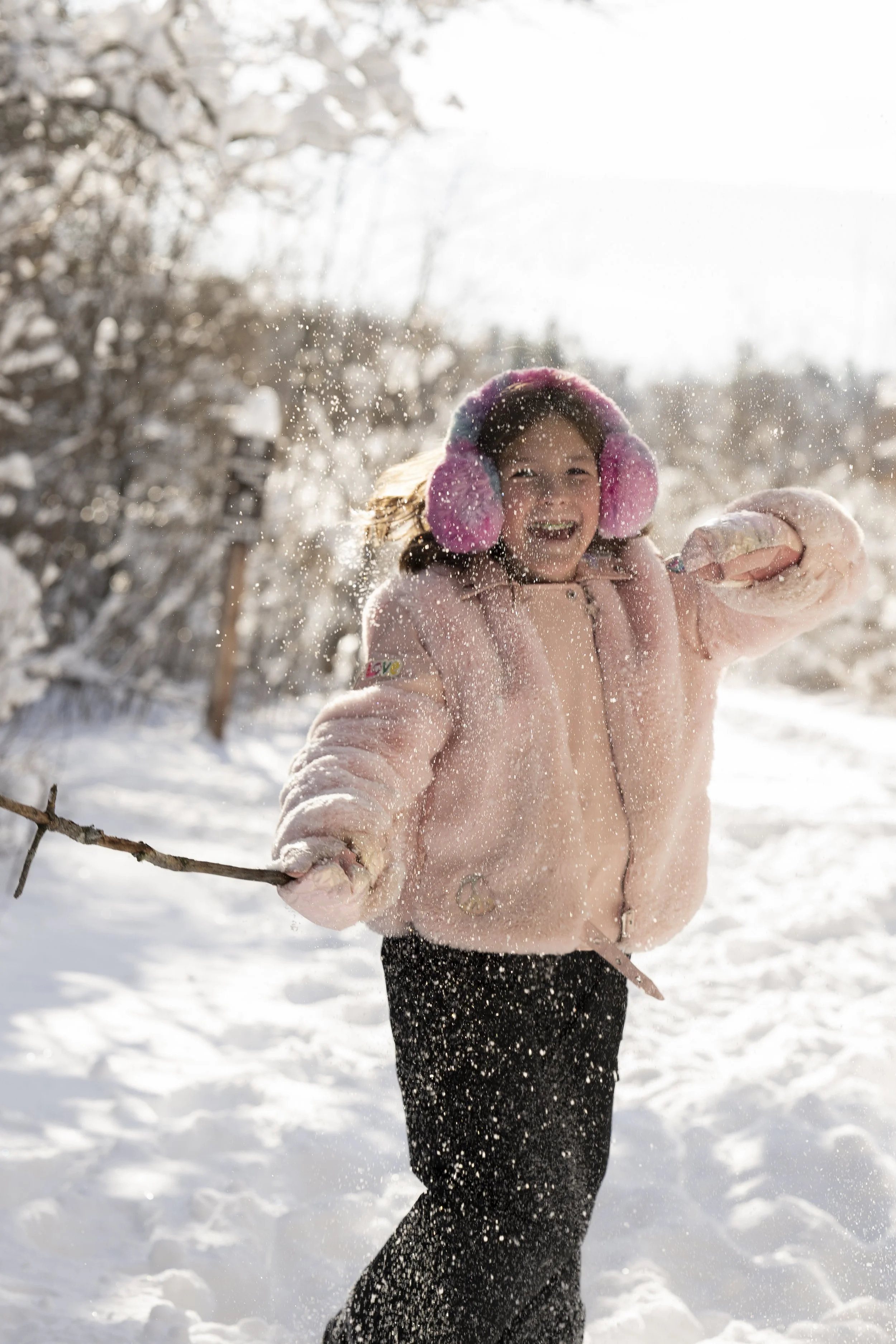 A young girl playing in the snow, smiling, wearing a pink winter coat, pink earmuffs, black pants, and a black hat, holding a stick, with snow around her and snow-covered trees in the background.