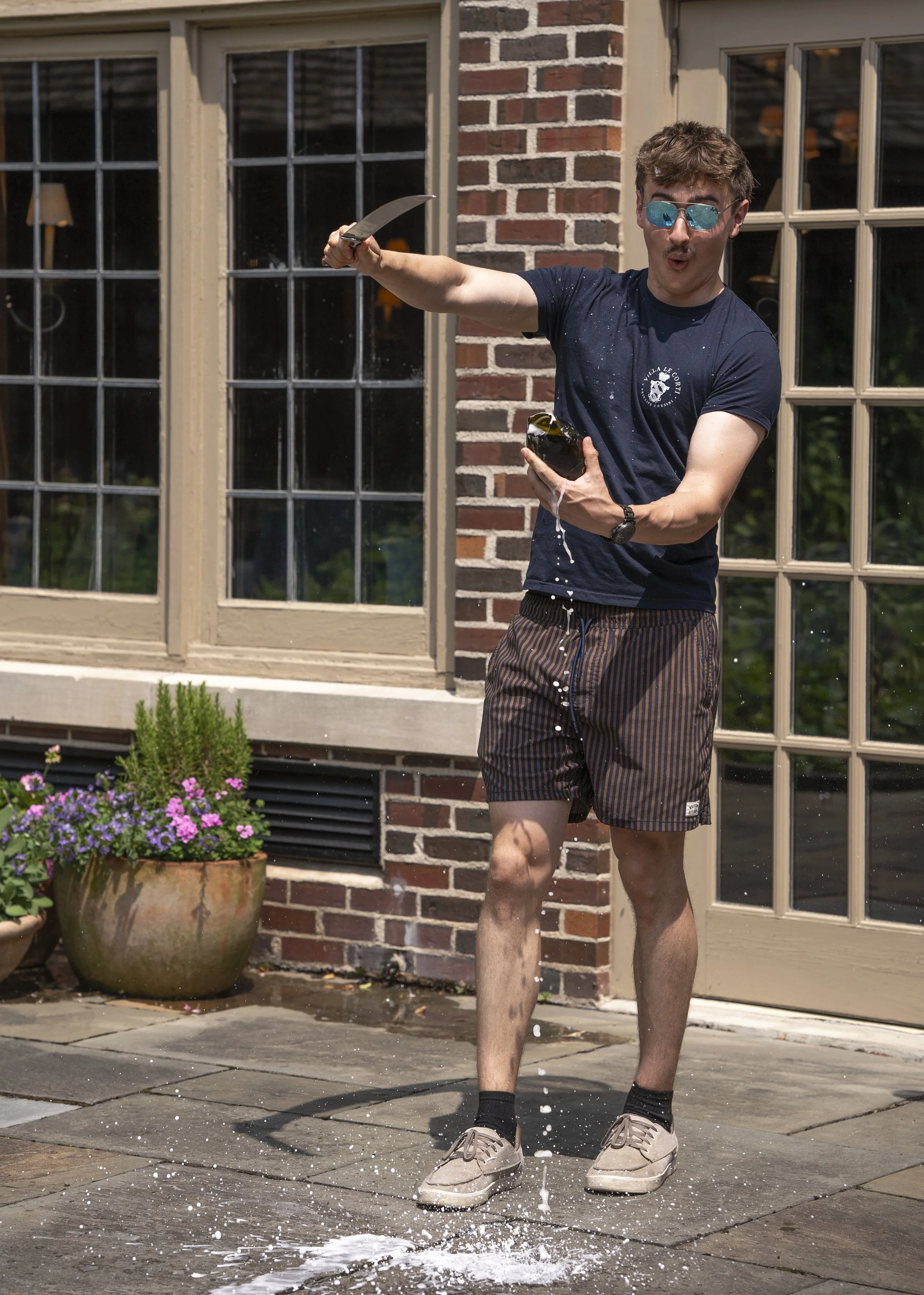 A young man in shorts and a t-shirt, wearing sunglasses, is standing outside on a concrete patio, splashing a bottle of champagne with water, with water spilling and splashing on the ground.