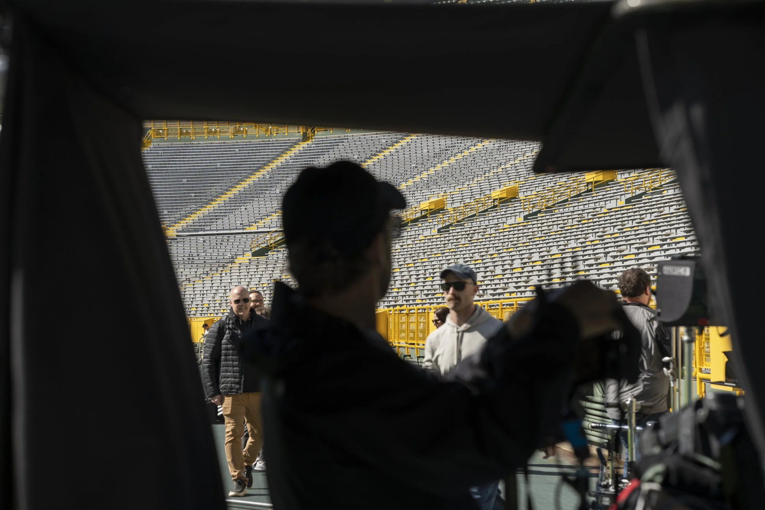 People on a sports field, seen through opening from inside a vehicle or equipment, with bleachers in the background.