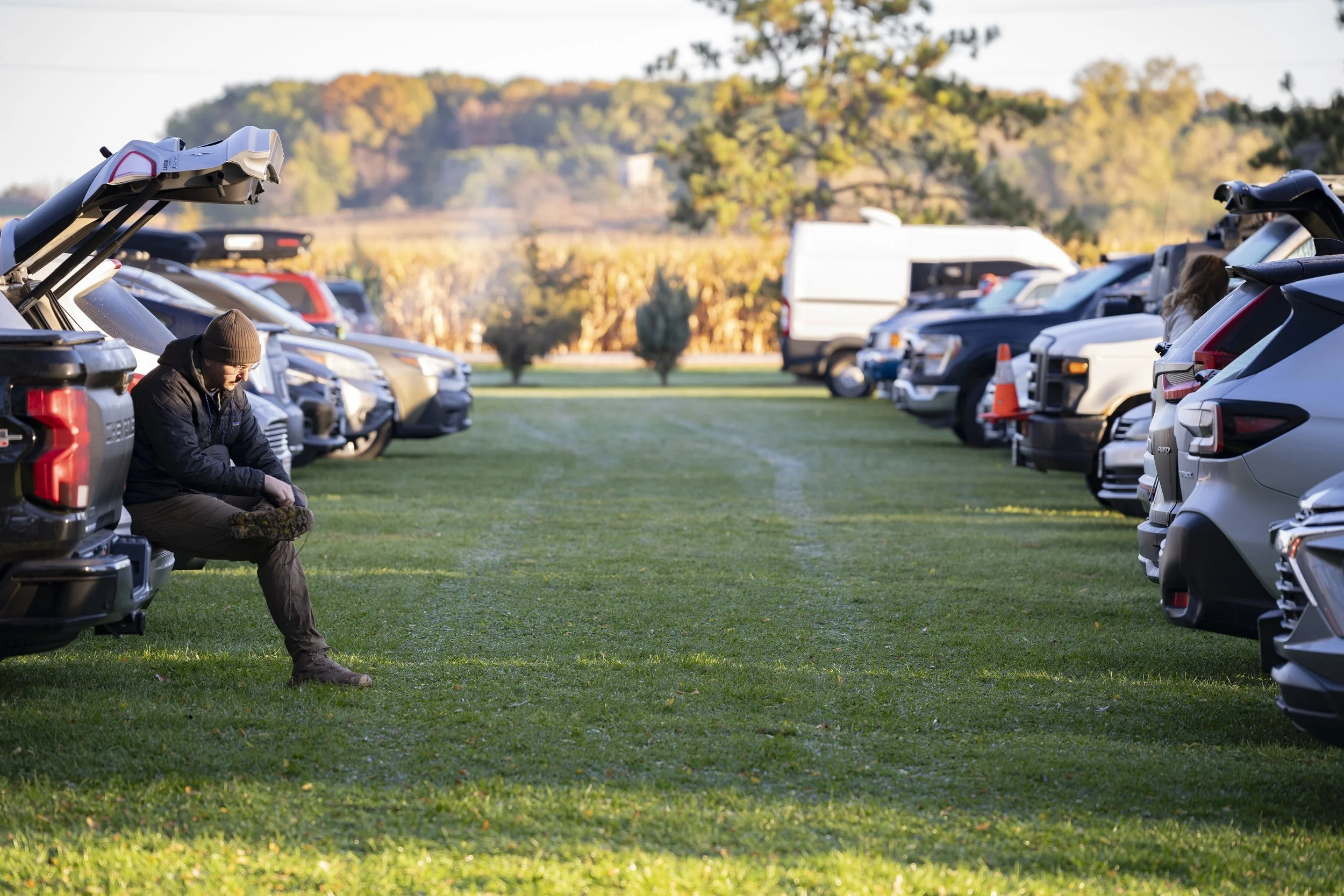 A man sitting on the open tailgate of a pickup truck in a park, with vehicles lined up on both sides and trees in the background.