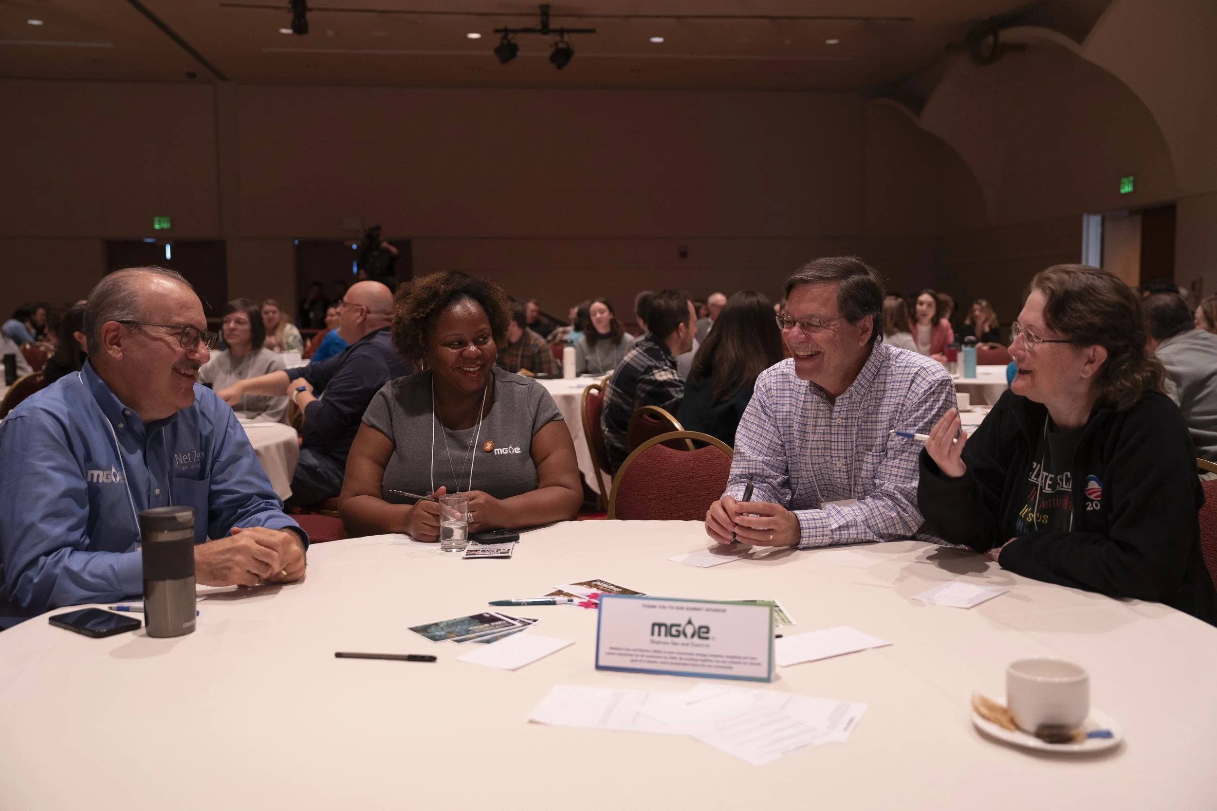 Group of four people sitting at a round table, engaged in conversation and smiling, at a conference or seminar in a large, dimly-lit room with many attendees.