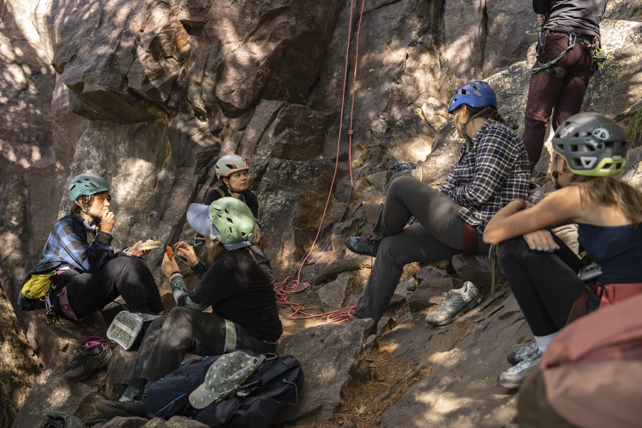 Group of six women wearing helmets, sitting and resting on rocks in a rocky outdoor setting during a rock climbing or rappelling trip.