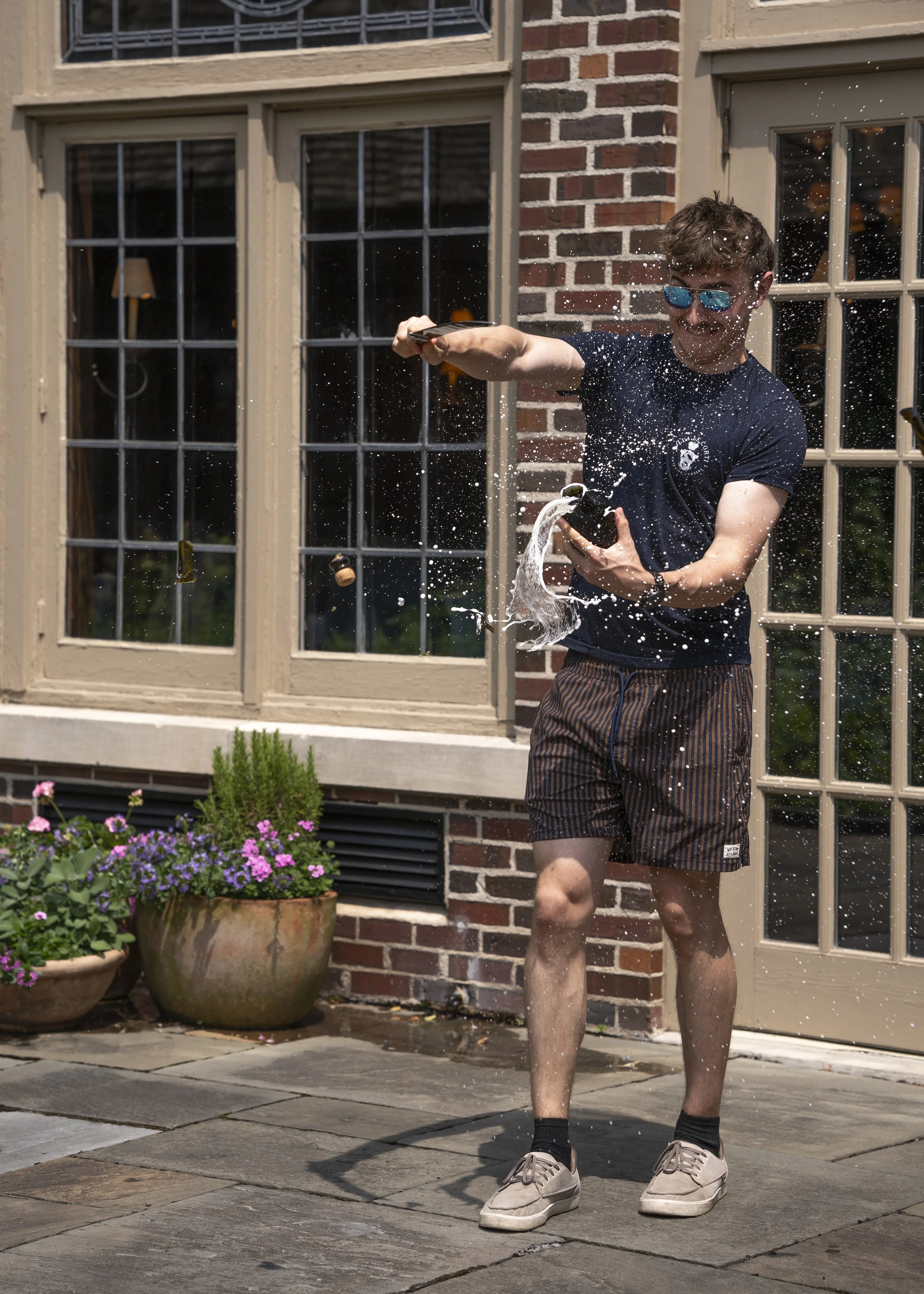A young man in sunglasses and casual clothes is outside, smiling and spraying a beverage or foam from a bottle, creating a splash of liquid.