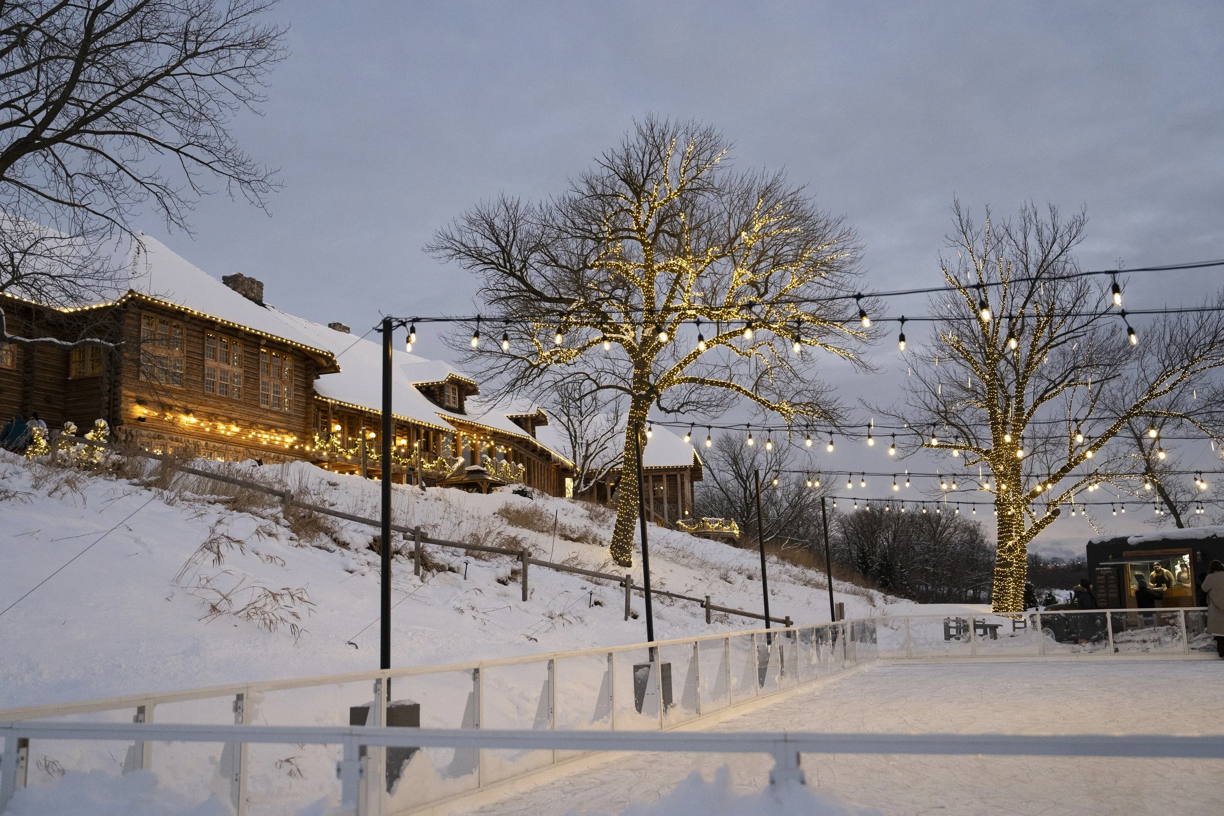Snowy outdoor ice skating rink at dusk with illuminated string lights and decorated trees, rustic wooden building in the background.