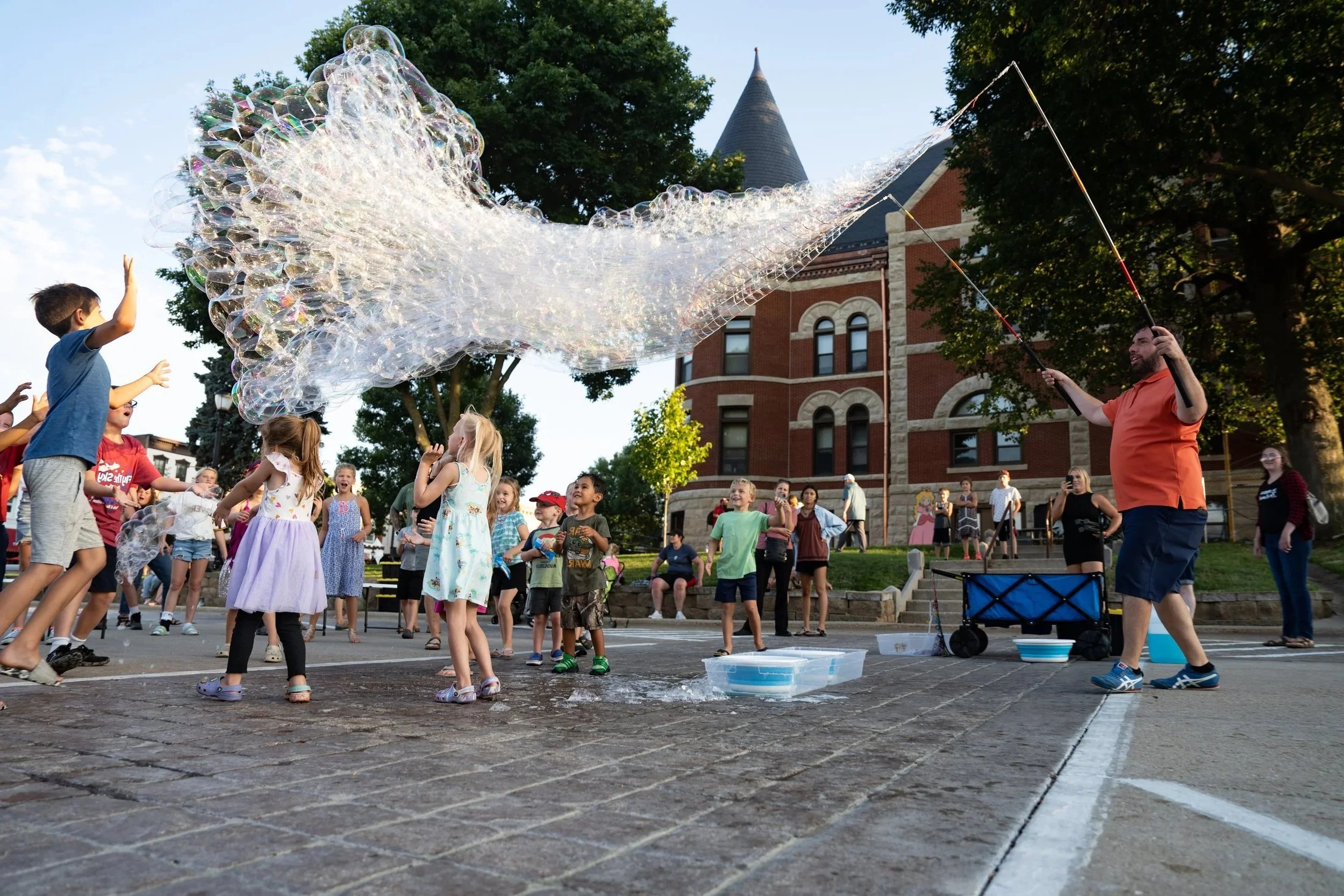 Children and adults gathered outside on a pavement, watching and playing with large soap bubbles created by a man with fishing poles, in front of a brick building with trees and a clear sky.