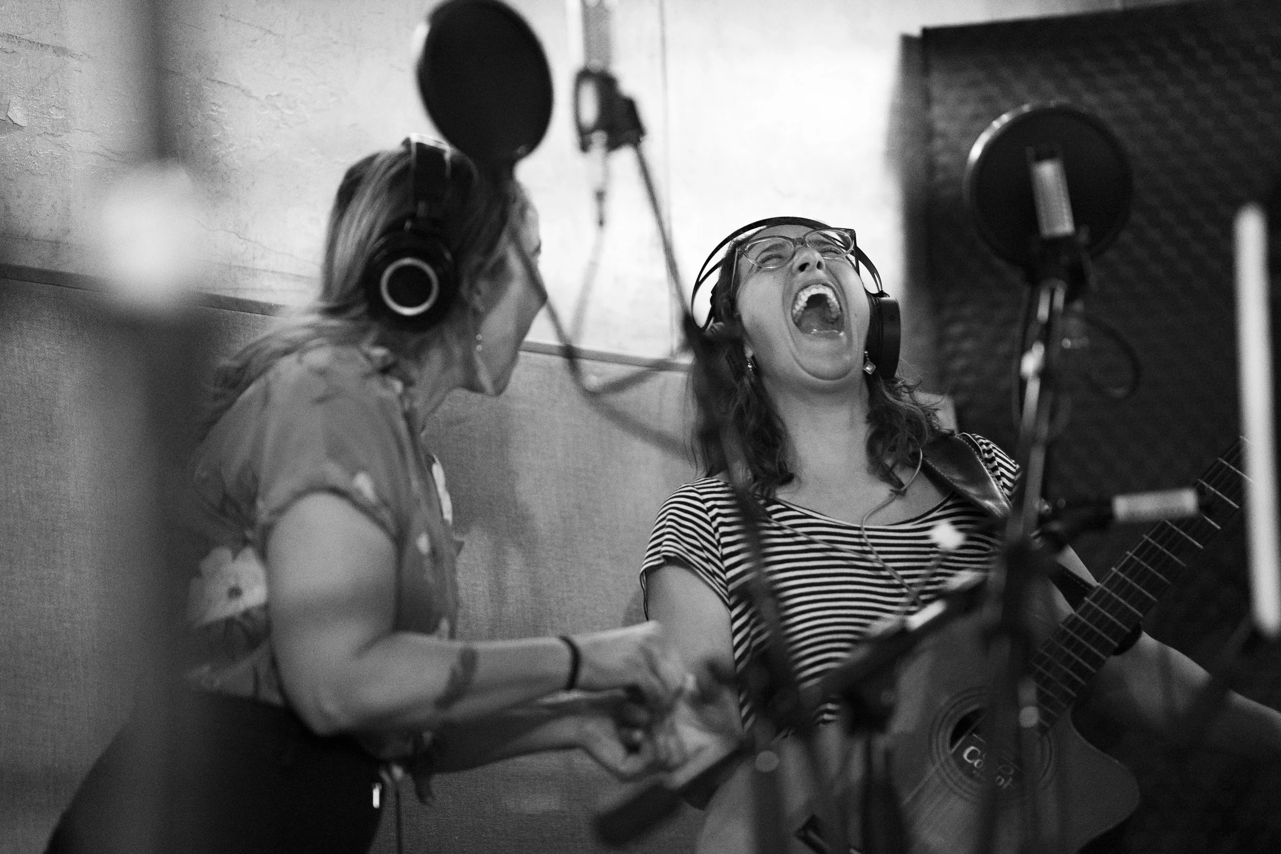 Two women in a recording studio, wearing headphones. One woman is singing passionately with her mouth wide open while playing a guitar. The other woman is holding a microphone laughing.