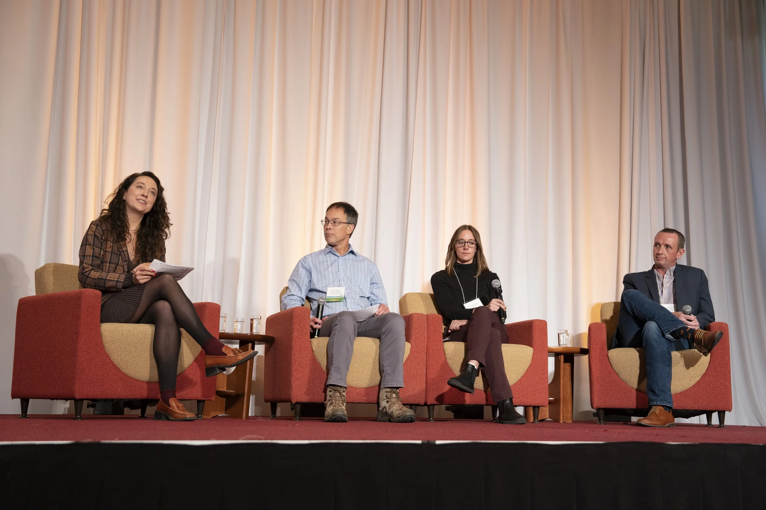 Four people sitting on stage in chairs, participating in a panel discussion with a woman on the far left holding notes and the other three holding microphones, in front of a cream-colored curtain.