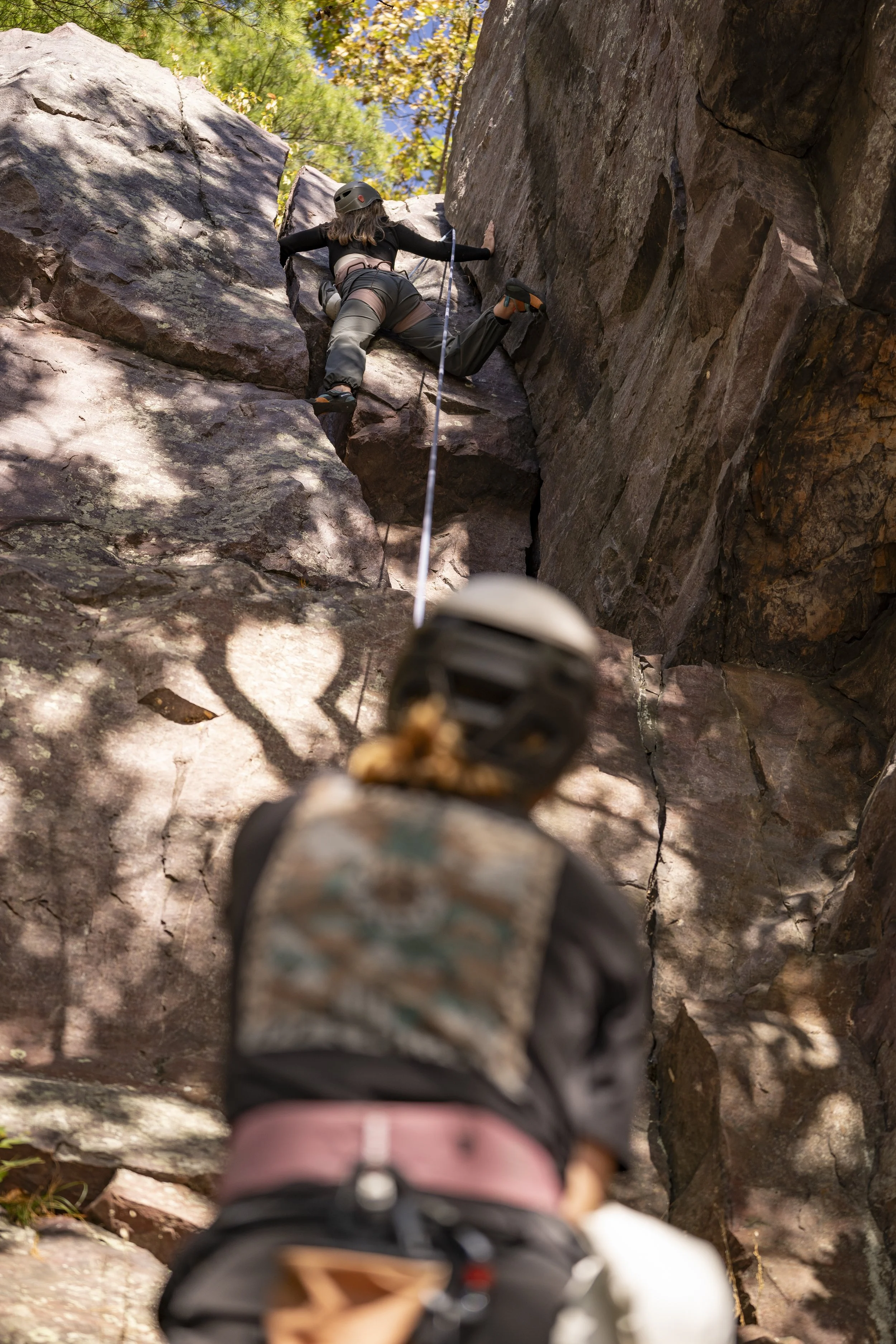 A woman climbing a steep rock face while being belayed by a person below, outdoors with trees and sunlight.