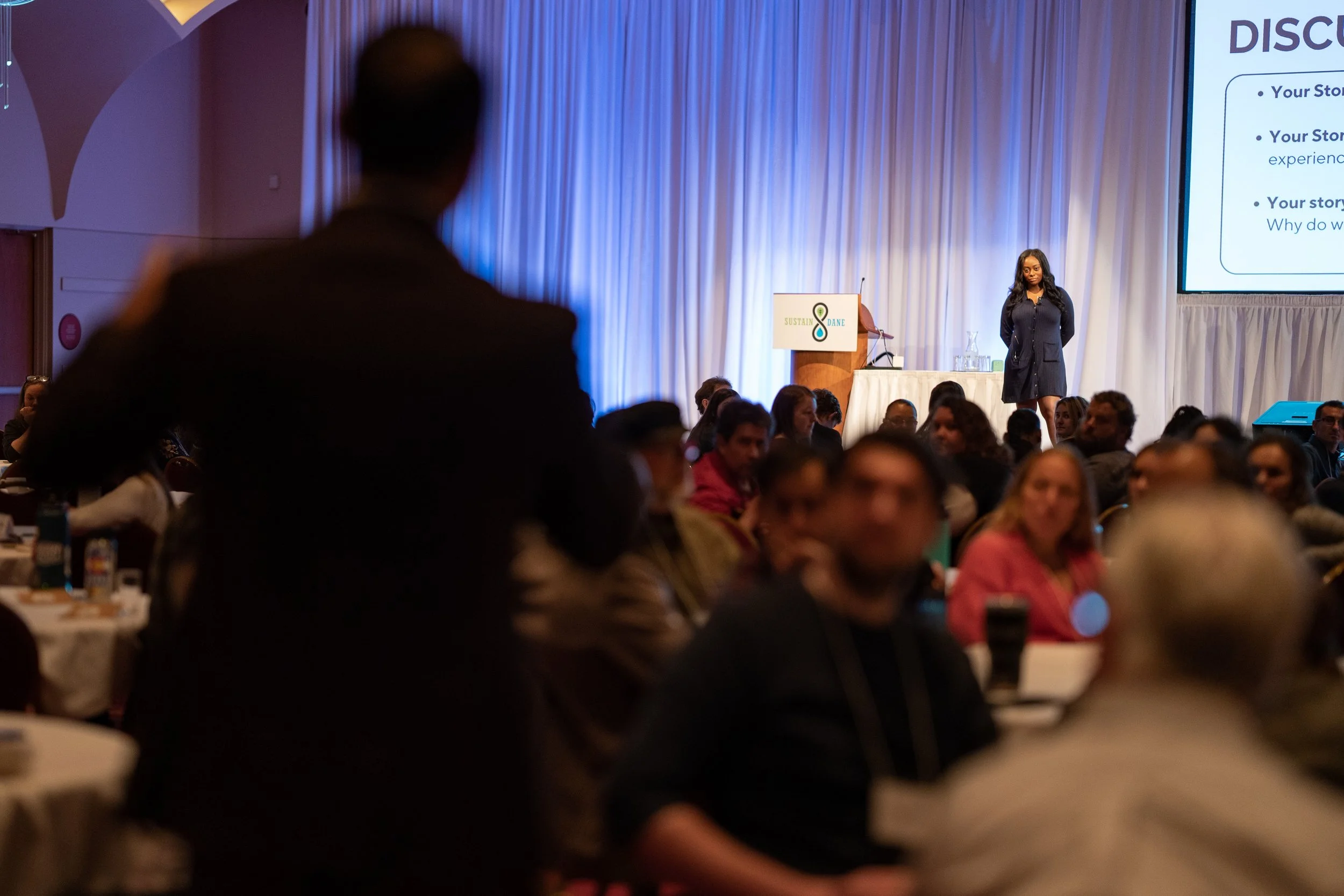 A conference or seminar with a woman on stage presenting, audience members seated at round tables, some looking towards the stage, and a large screen displaying presentation slides.