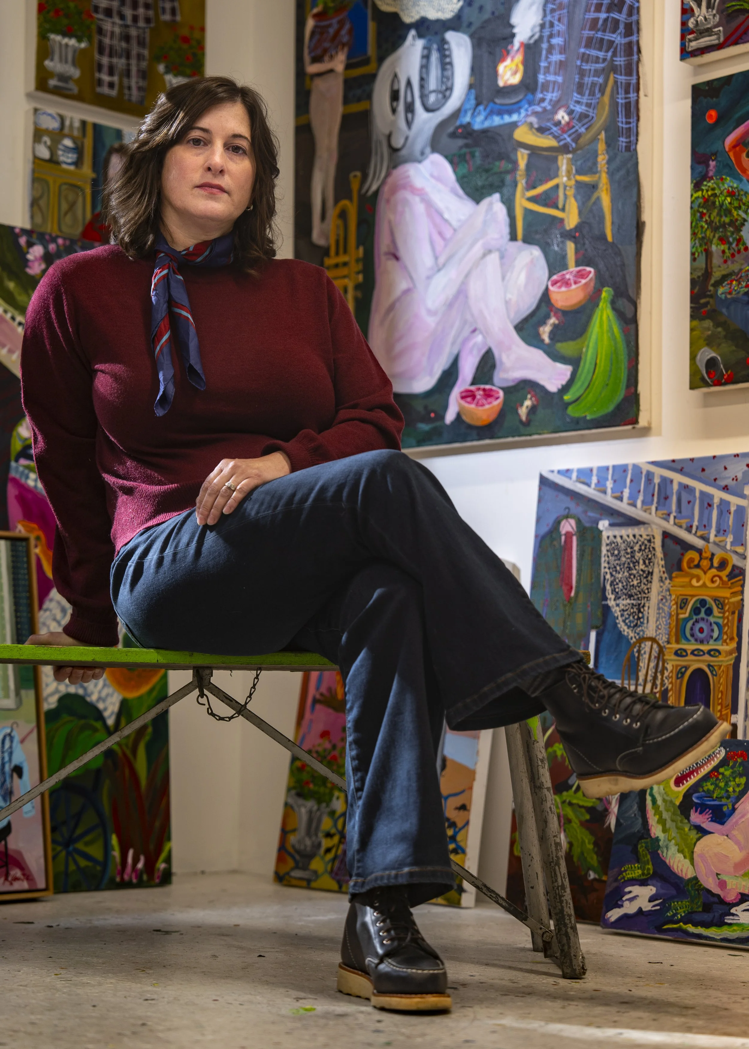 A woman with shoulder-length dark hair, wearing a red sweater, blue jeans, black boots, and a navy scarf with red stripes, sitting on a green platform in front of colorful paintings, including a large artwork featuring a skull, a figure with crossed 