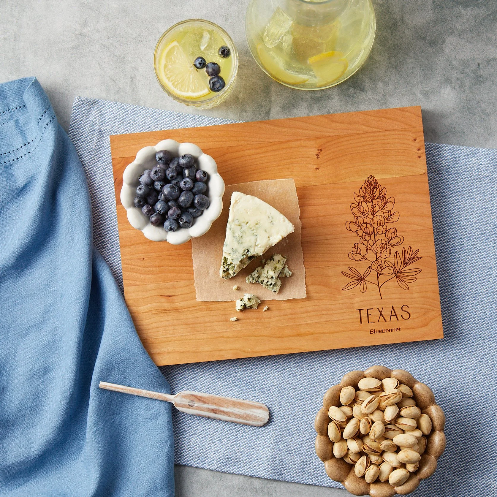 A Texas state flower inscribed cutting board styled with some blueberries, blue napkins, blue cheese, pistachios and lemonade. 