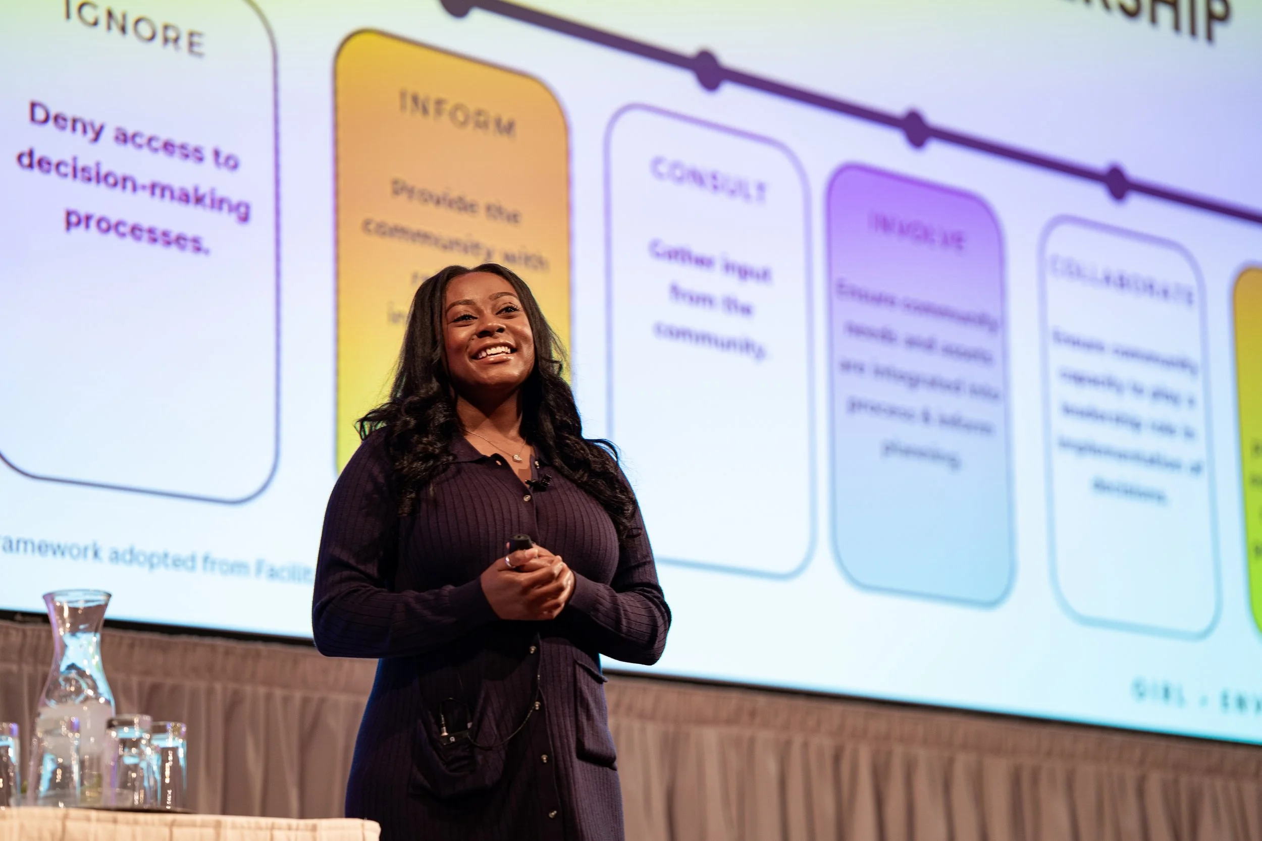 A woman standing in front of a large presentation slide, smiling with her hands clasped.