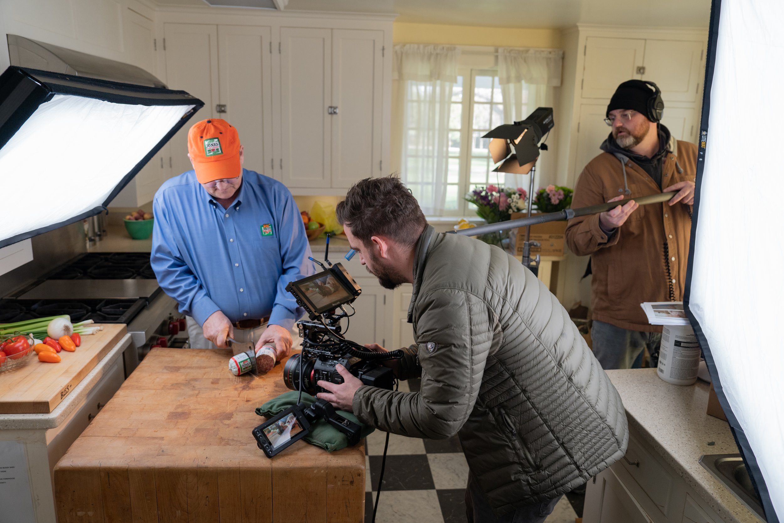 Behind the scenes of a video shoot in a kitchen, with a man filming while another prepares ingredients, and a crew member adjusting lighting.
