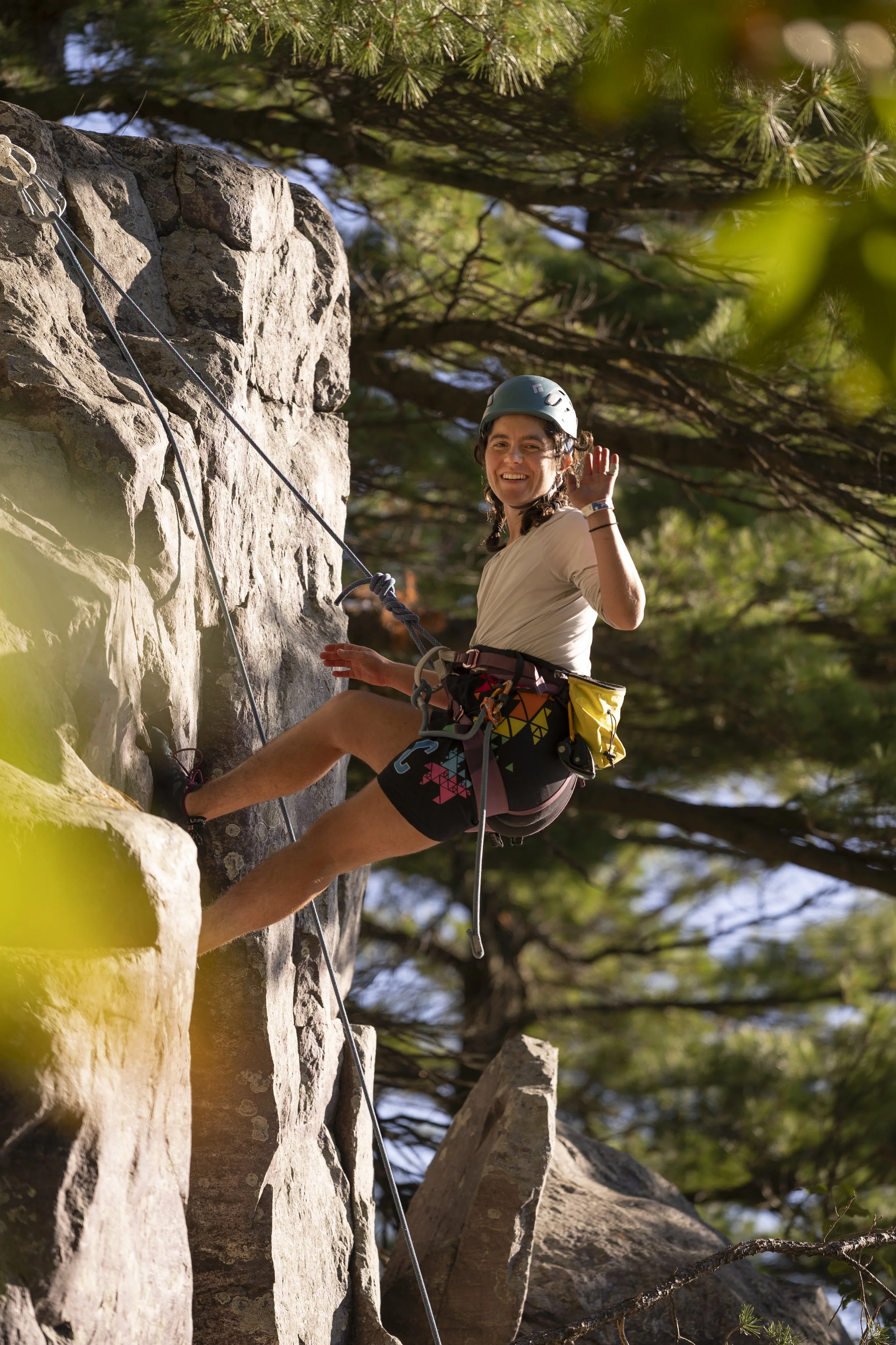 A woman rock climbing outdoors, wearing a helmet and smiling, while waving to the camera.