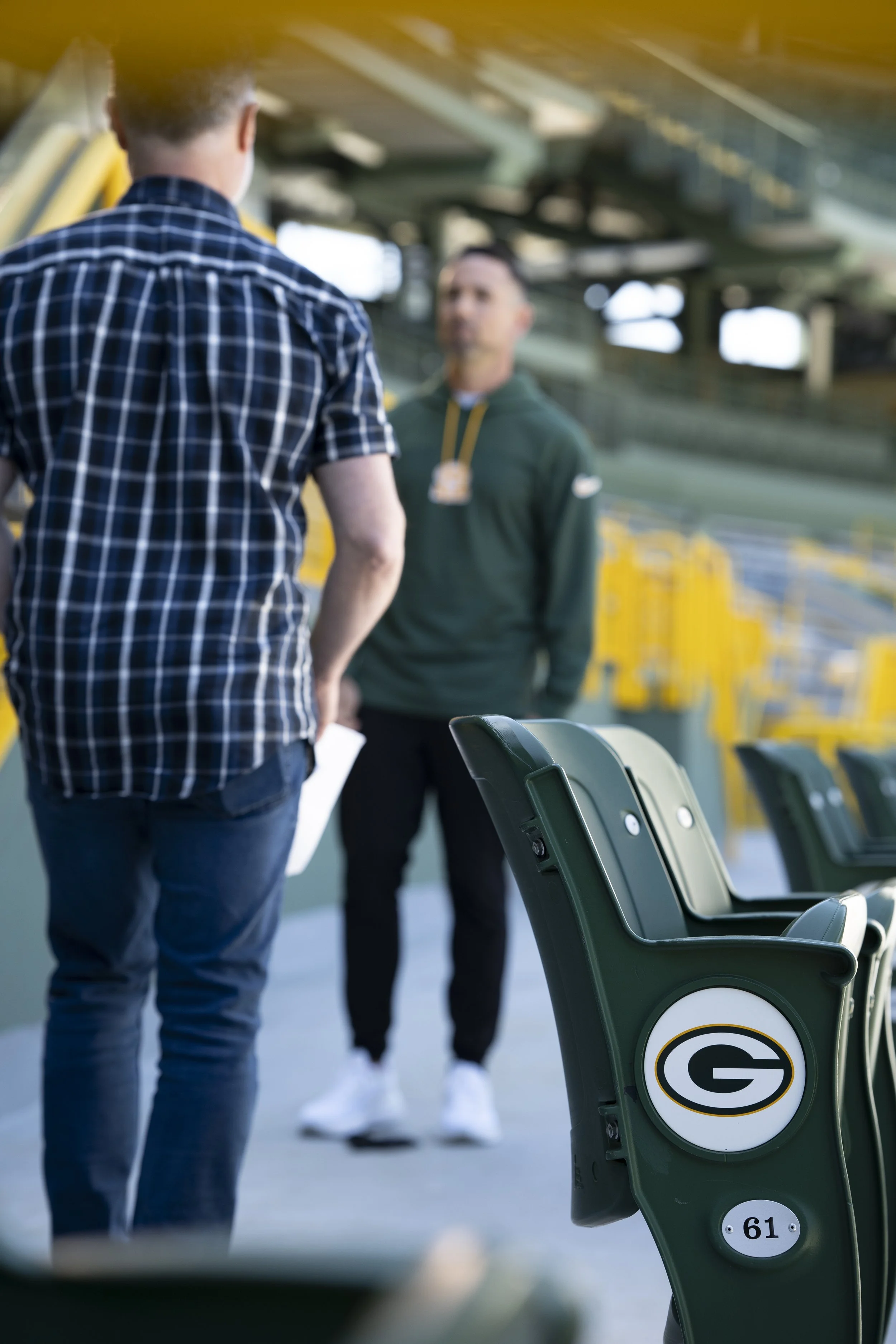 Two men having a conversation at a football stadium, one facing away and wearing a checkered shirt, the other facing forward in a green hoodie, with stadium seats and yellow structures in the background.