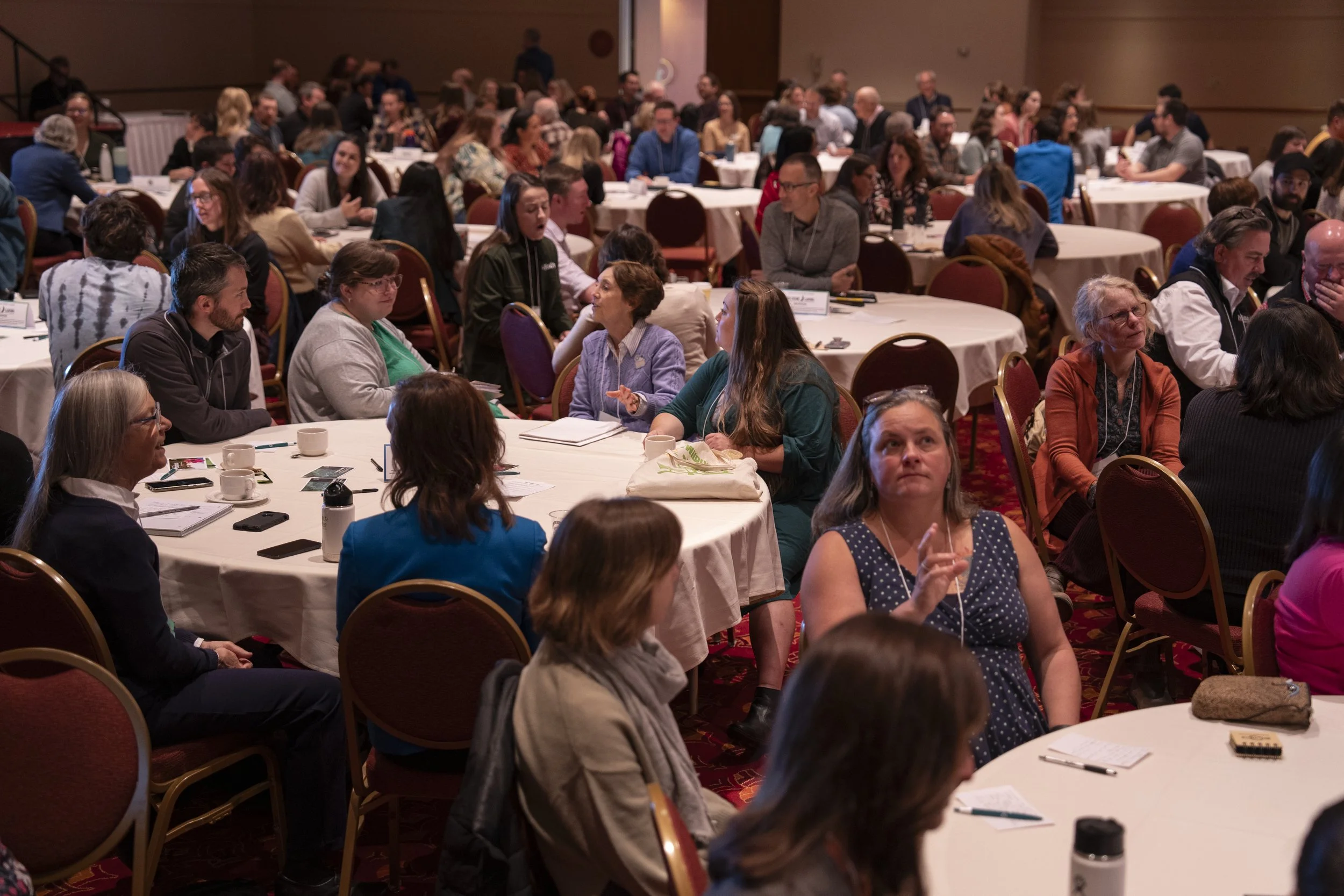People sitting at round tables in a conference room, engaged in conversations and listening to a speaker, with some taking notes.