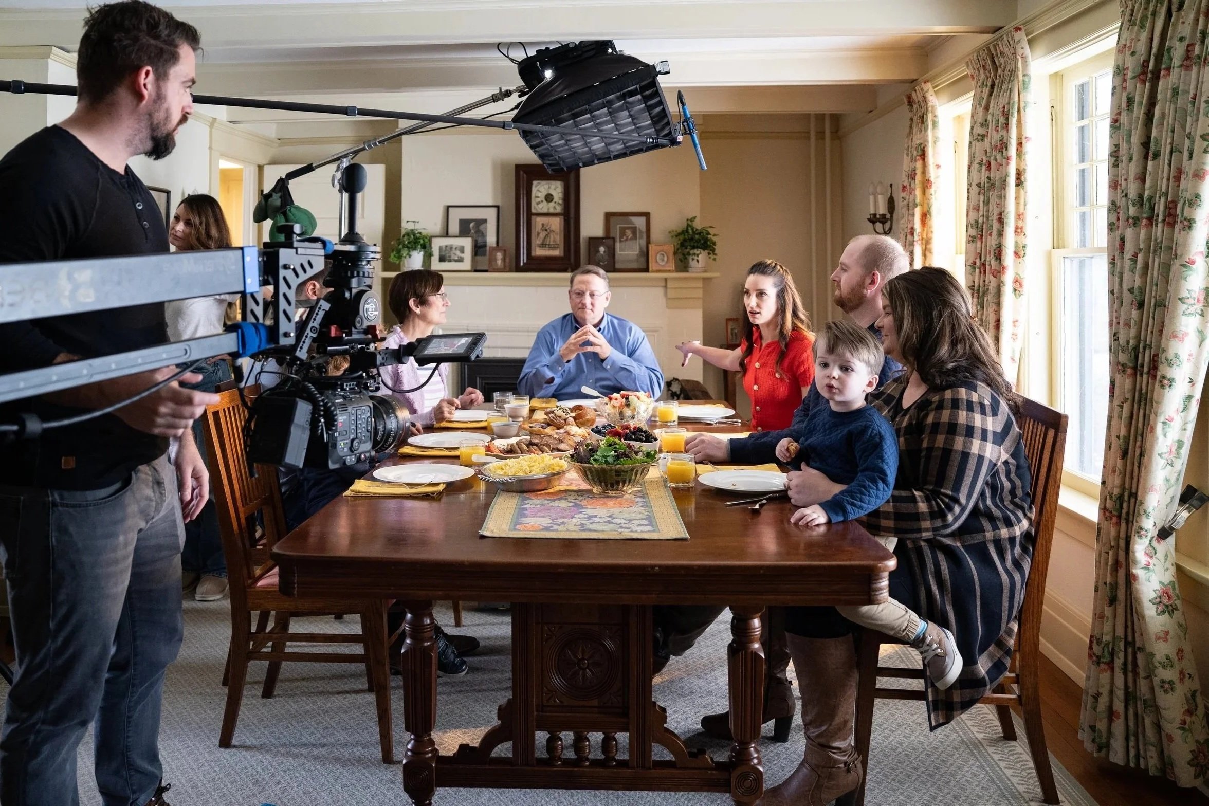 Film crew filming a family breakfast at a dining table in a bright, cozy room with floral curtains and decorative pictures on the wall.