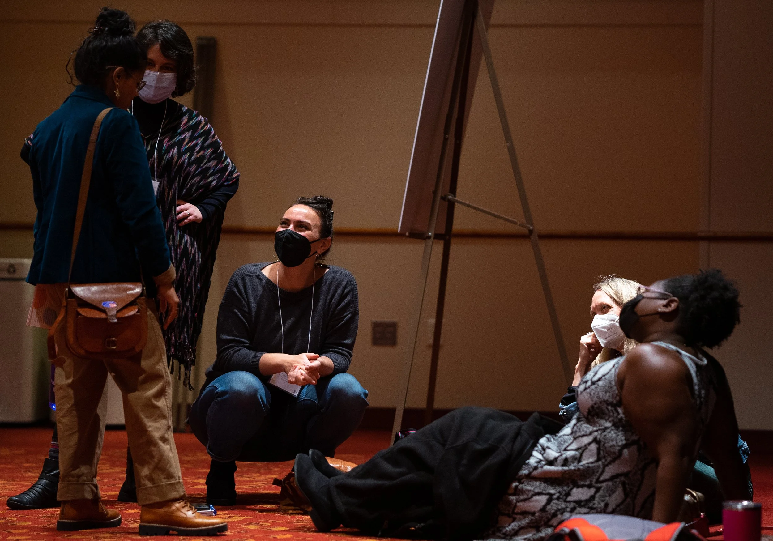 Group of five women, four sitting on the floor and one standing, engaged in conversation at indoor event, all wearing face masks.
