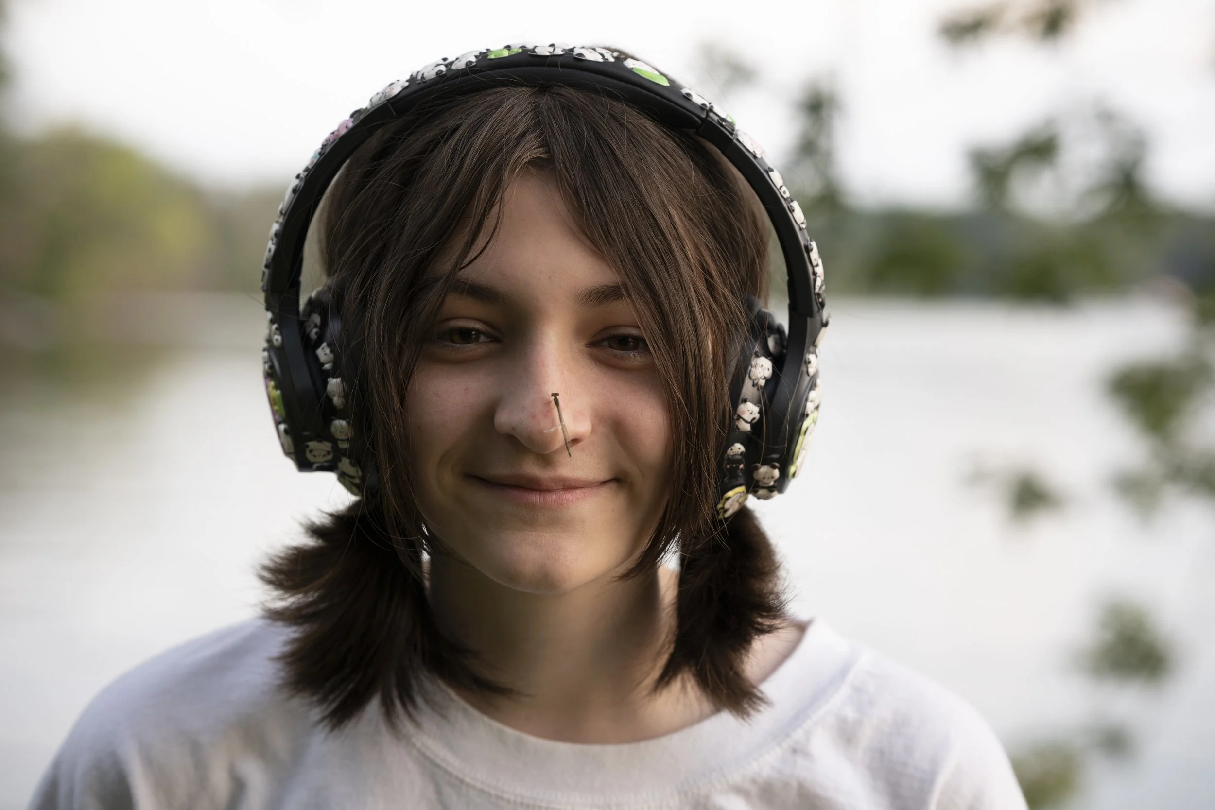 Young person outdoors wearing large headphones with cartoon character designs, smiling, with a body pin in her nose, and a lake and trees in the background.