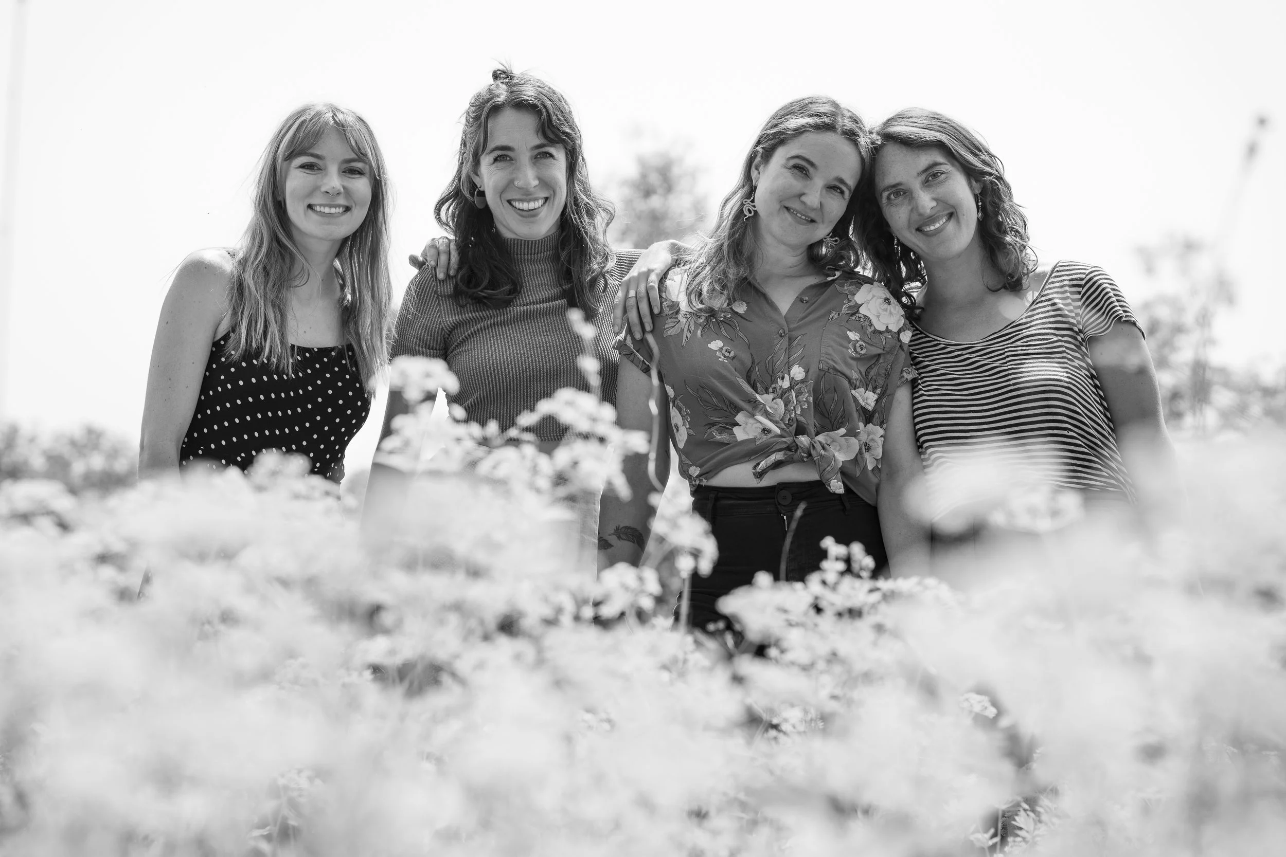 Black and white photo of five smiling women standing outdoors among flowers, with some women’s arms around each other.
