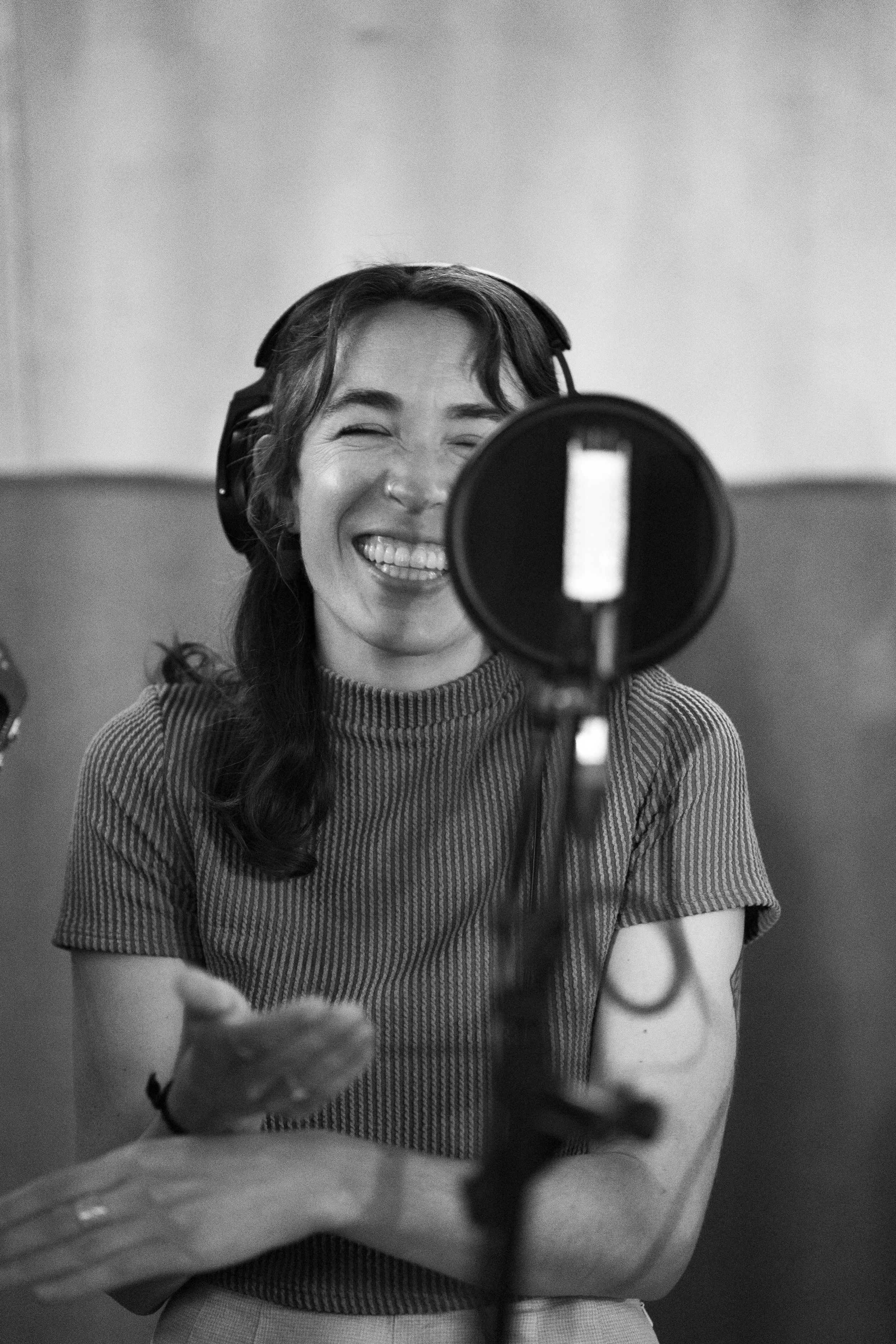 A woman in a recording studio smiling and wearing headphones, sitting in front of a microphone.