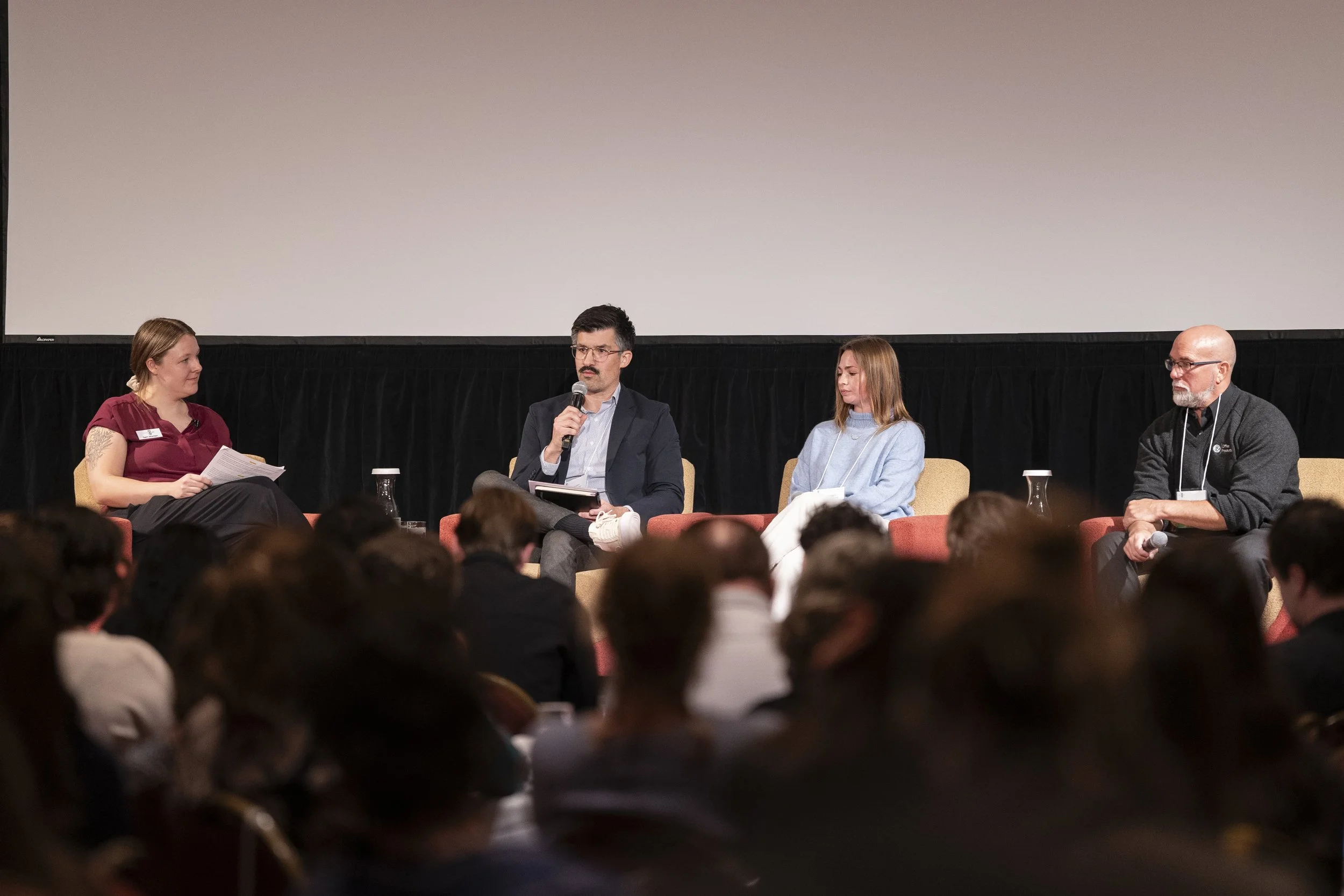 Four people sitting on a stage in front of an audience during a panel discussion. A woman on the left, a man with a microphone in his hand, a woman in a light blue sweater, and a man with glasses and a beard hold a microphone.