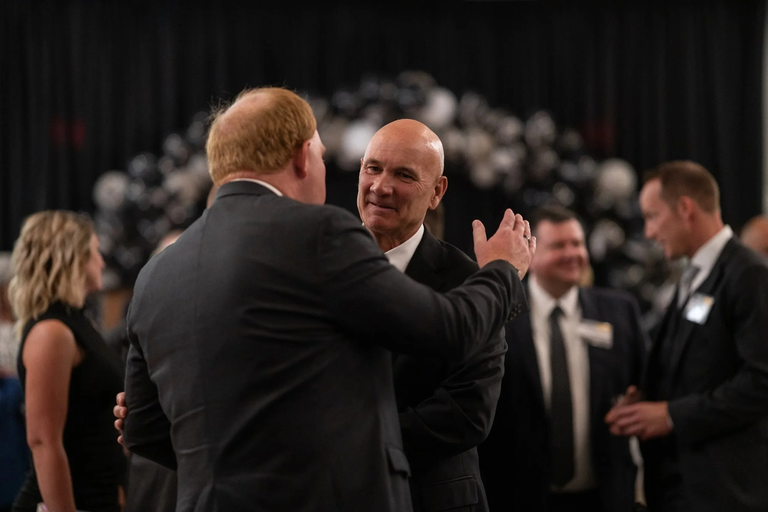 Two men in suits having a conversation at a formal event with other people in the background.