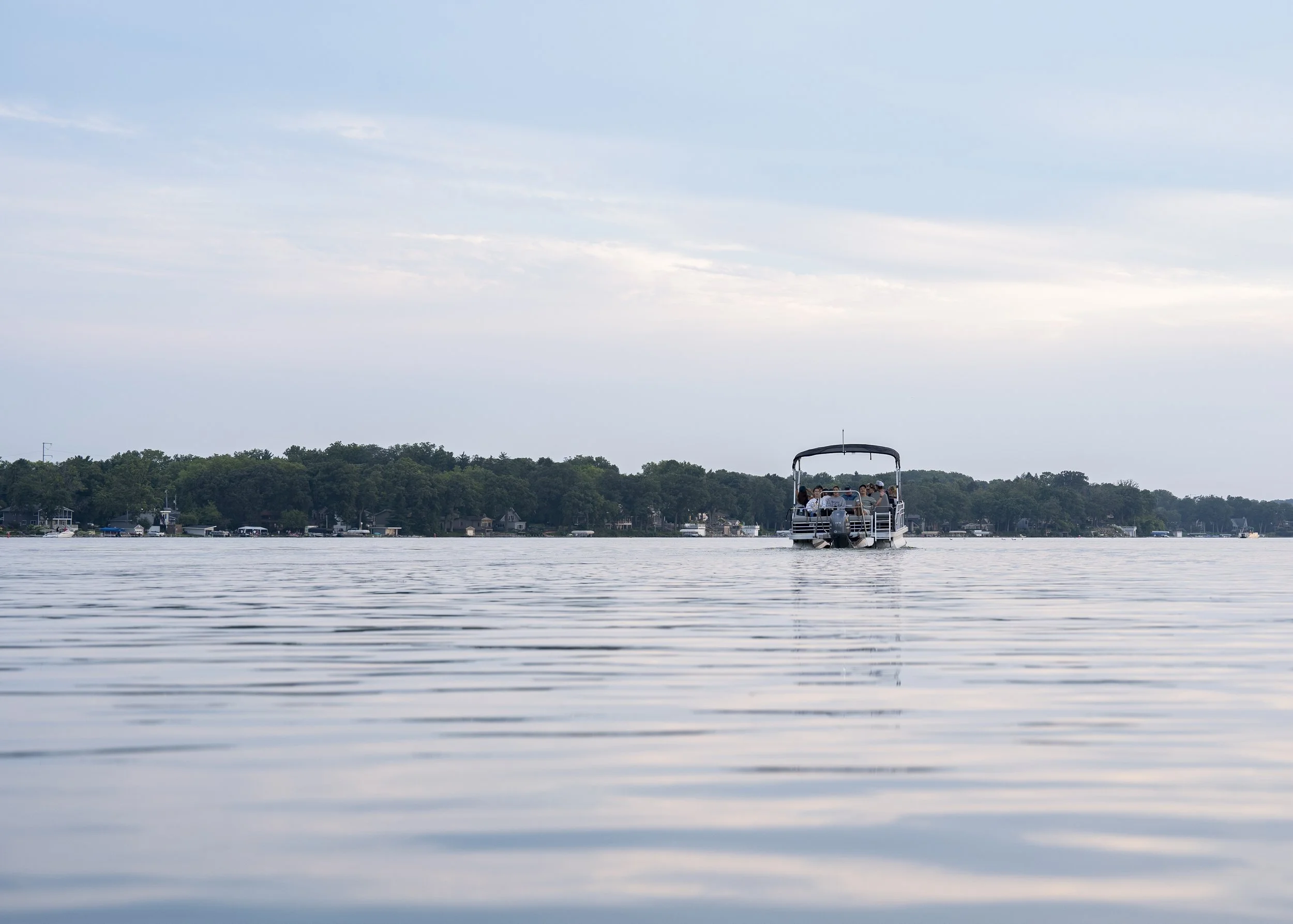 A boat with passengers on a calm lake, with houses and trees in the background under a cloudy sky.