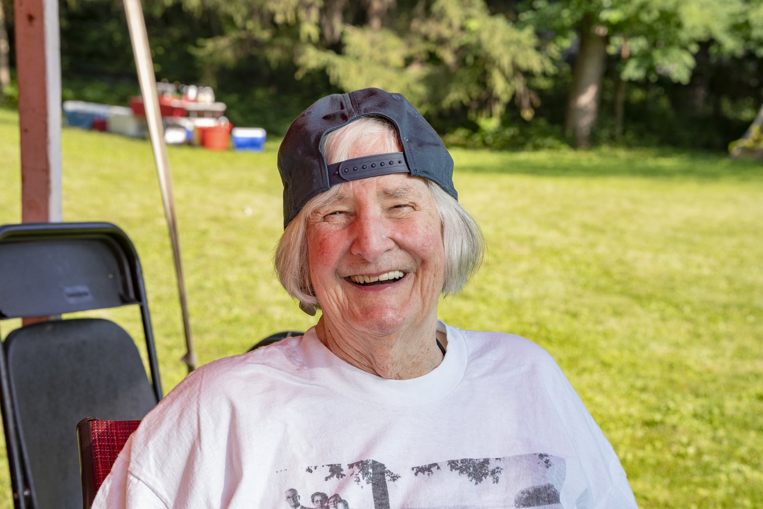 An elderly woman with gray hair, wearing a white t-shirt and a backward navy blue baseball cap, is smiling outdoors on a sunny day. There are trees and a grassy area in the background.