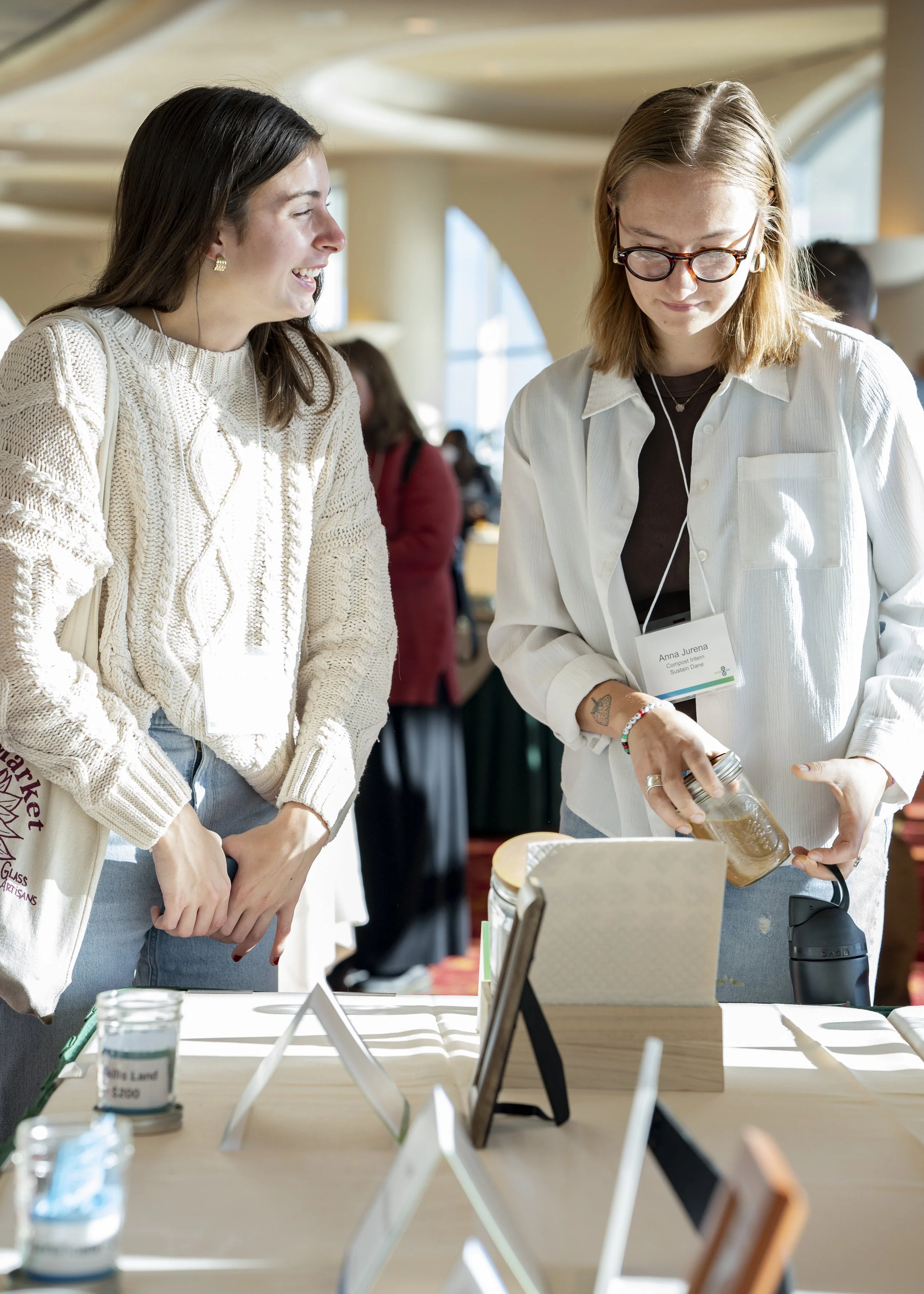 Two women at a table with jars and display signs in a well-lit indoor space. One is laughing and the other is putting paper in a jar. 
