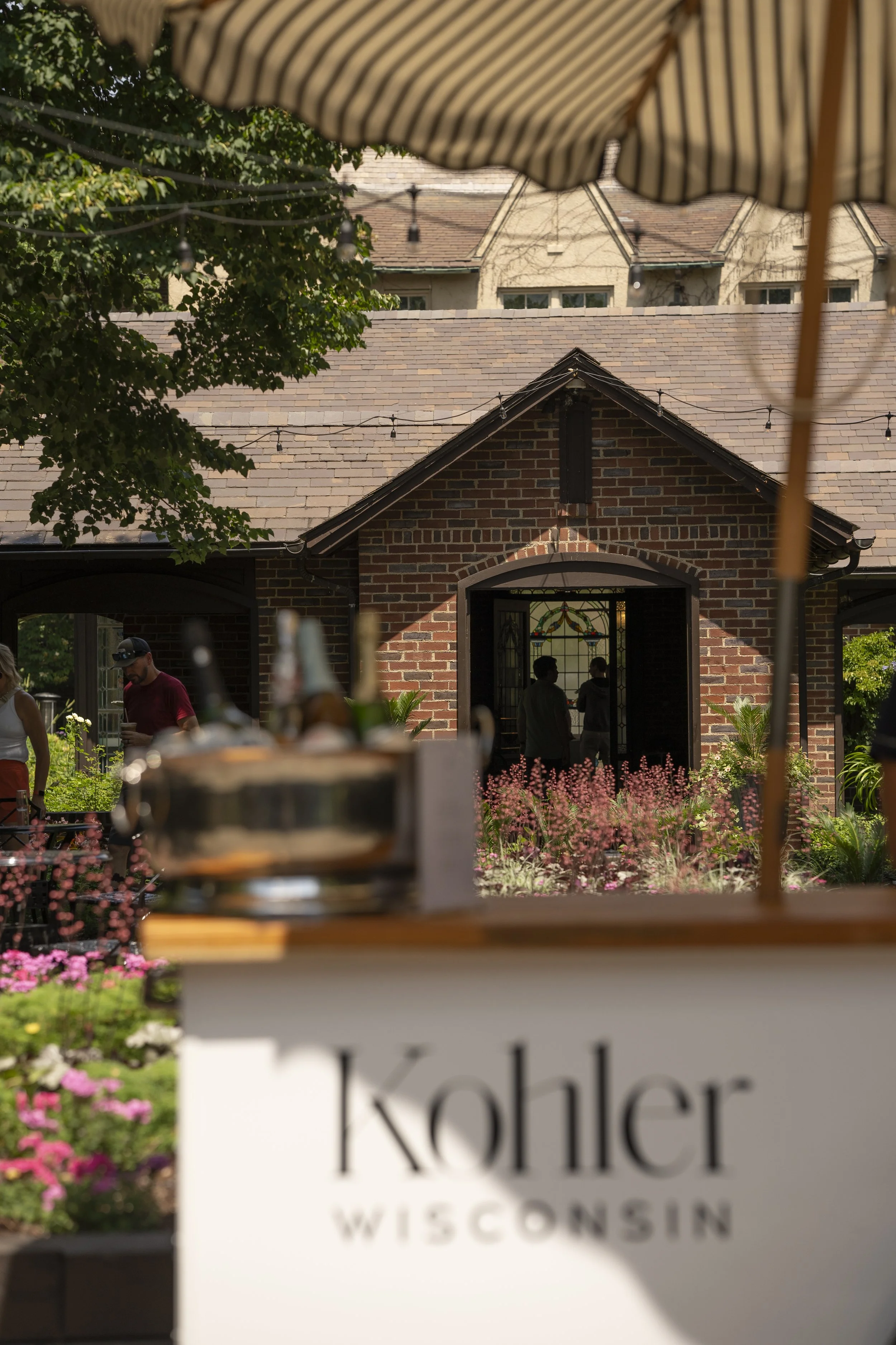 A scene of an outdoor gathering at Kohler, Wisconsin, with a brick building and people in the background. The foreground includes a blurry sign with the word 'Kohler' and some tables and flowers under a striped umbrella.