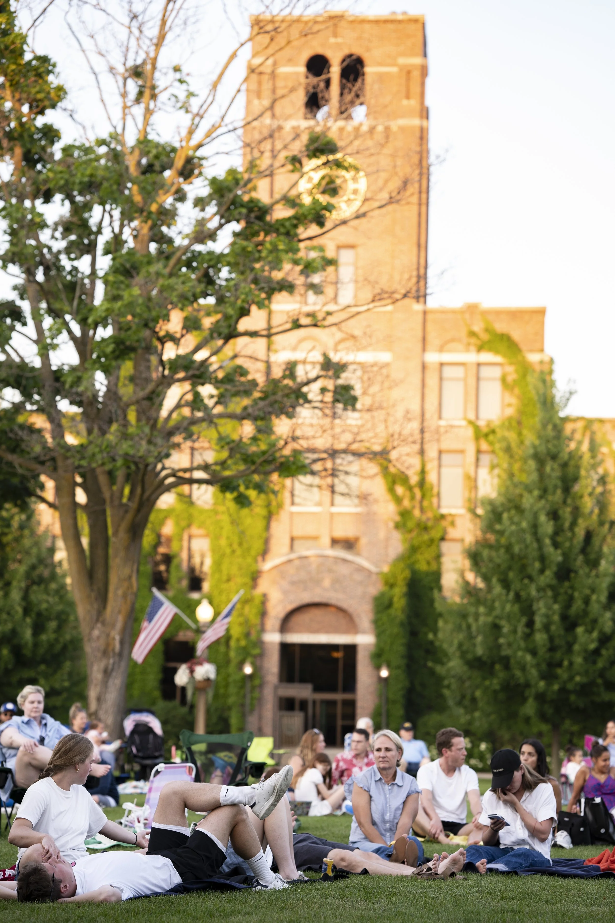 People sitting and lying on the grass in front of a historic brick building with a tower, during a community event or gathering.