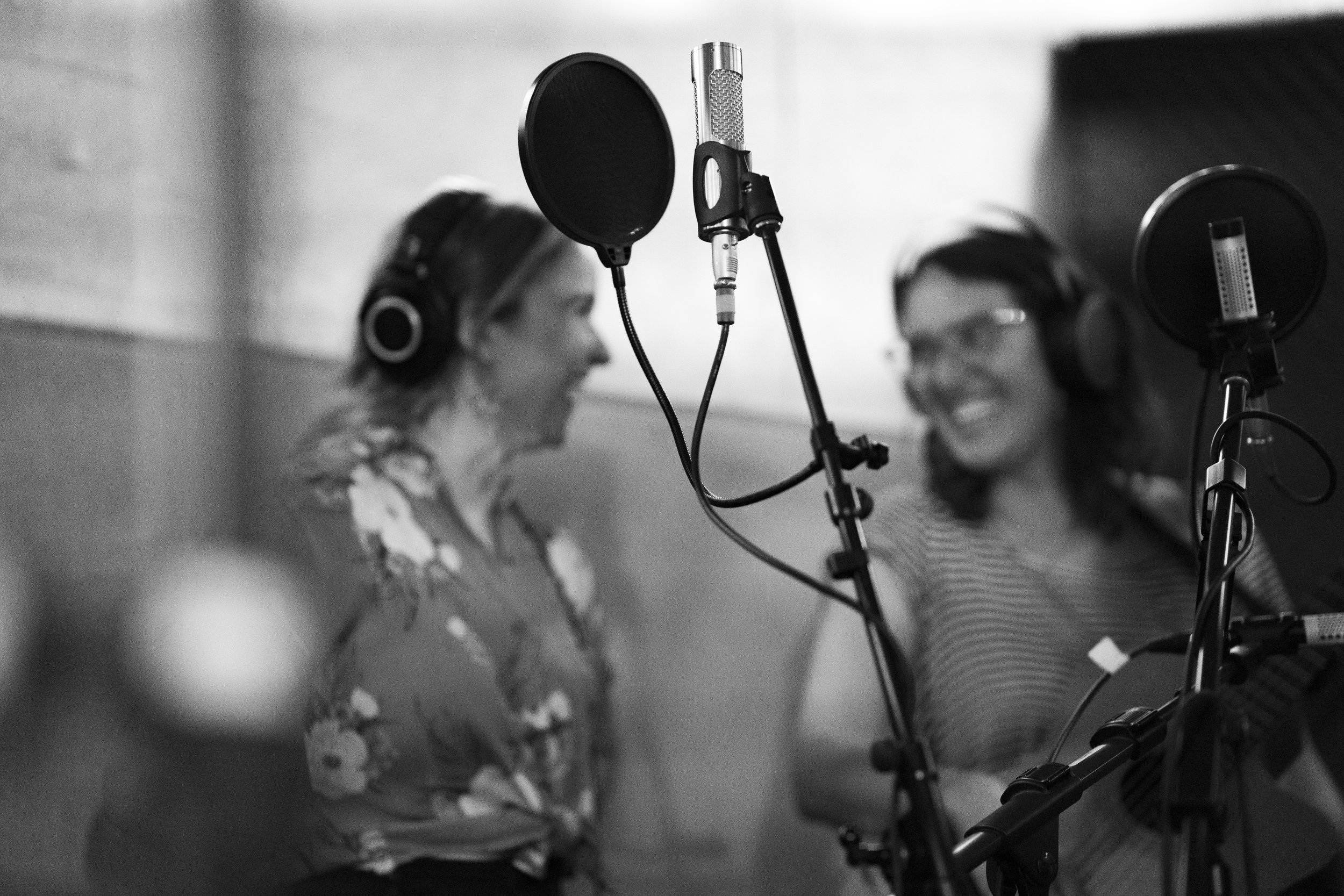 Two women in a recording studio wearing headphones, singing into separate microphones, and smiling at each other.