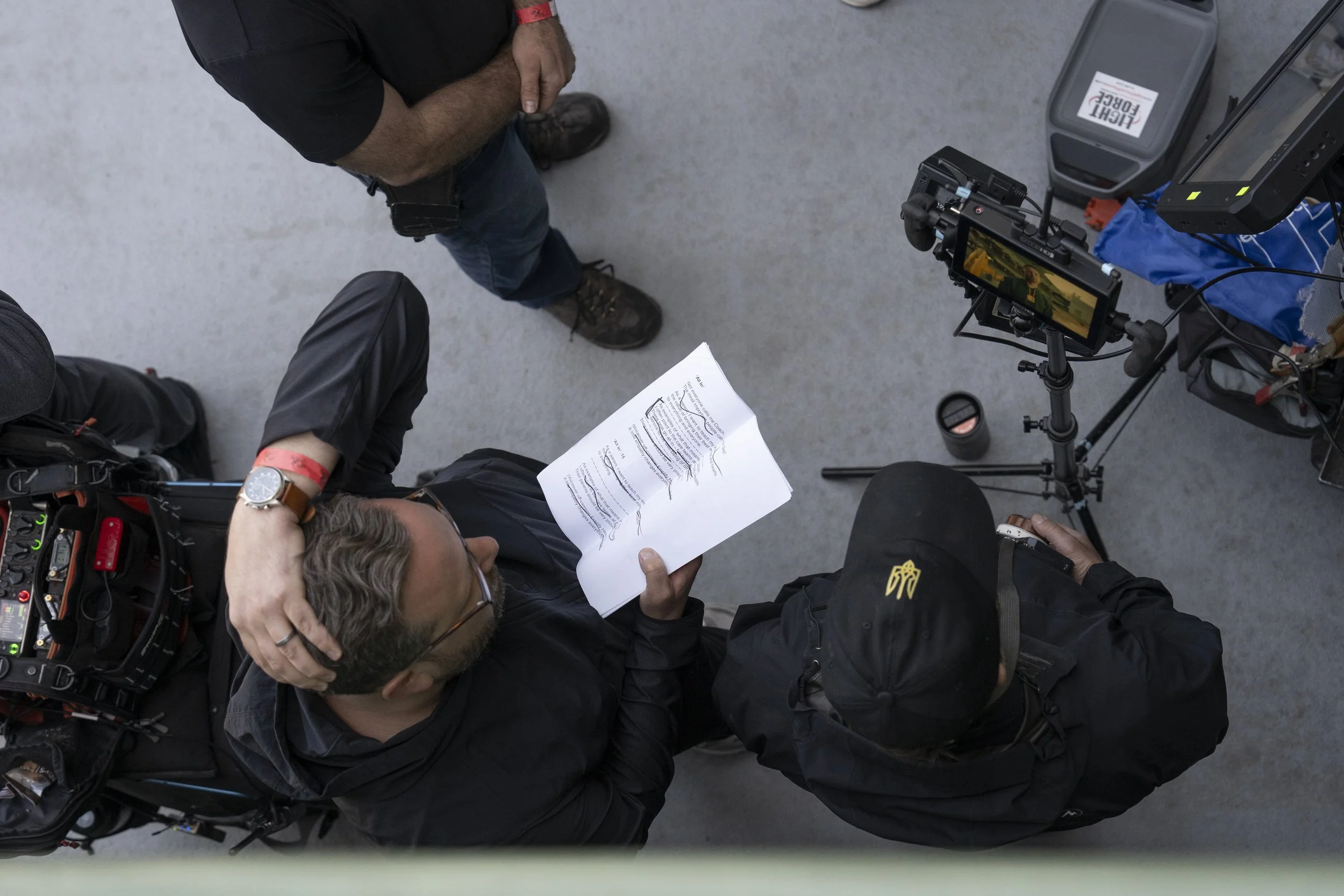 Two men engaged in conversation on a film or photography set, with filming equipment such as cameras, tripods, and a monitor around them. One man is reading a script, while the other is talking and wearing a black cap.