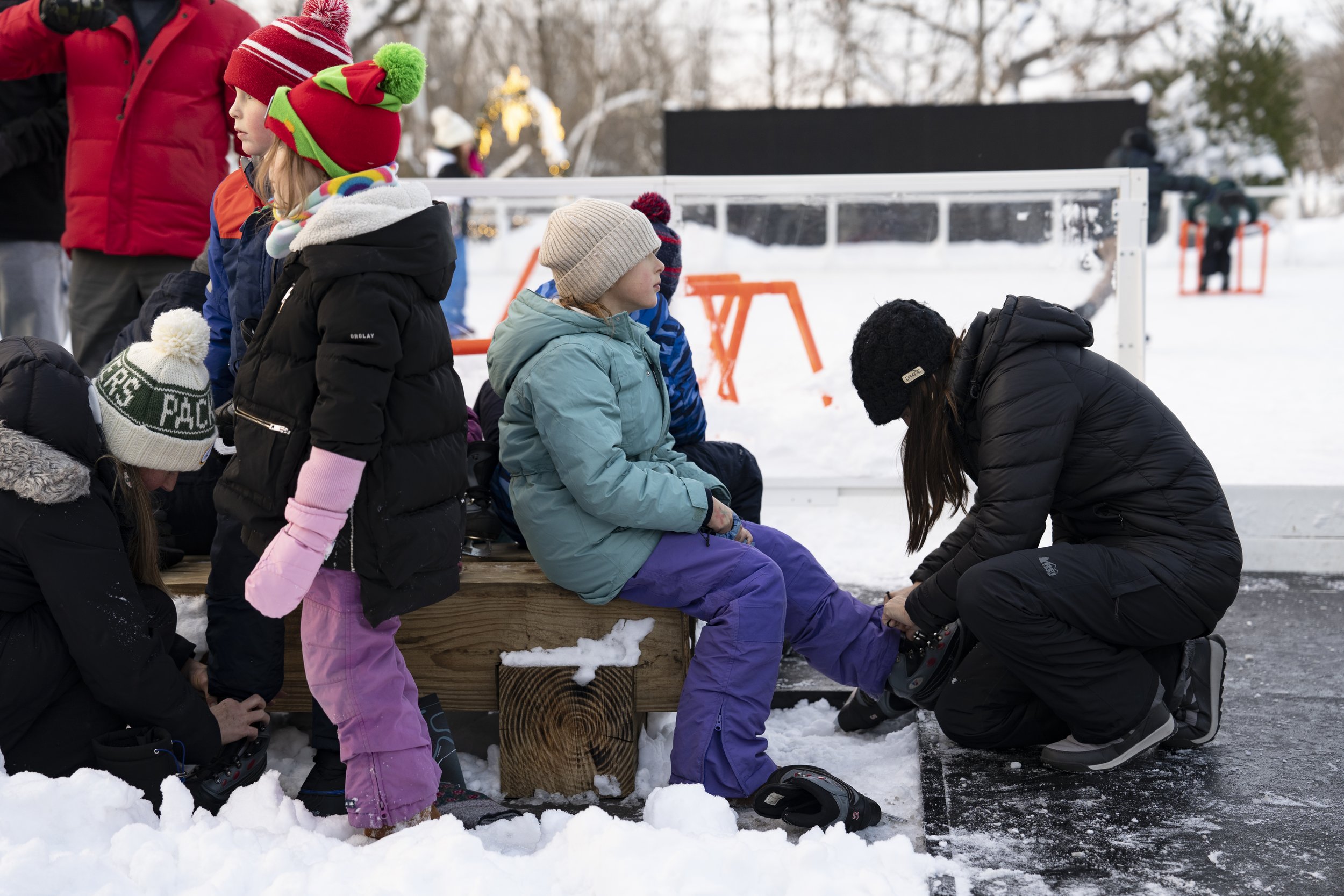 A group of children dressed in winter clothing, sitting and standing on a snow-covered surface near an ice skating rink, as an adult helps a girl put on ice skates.