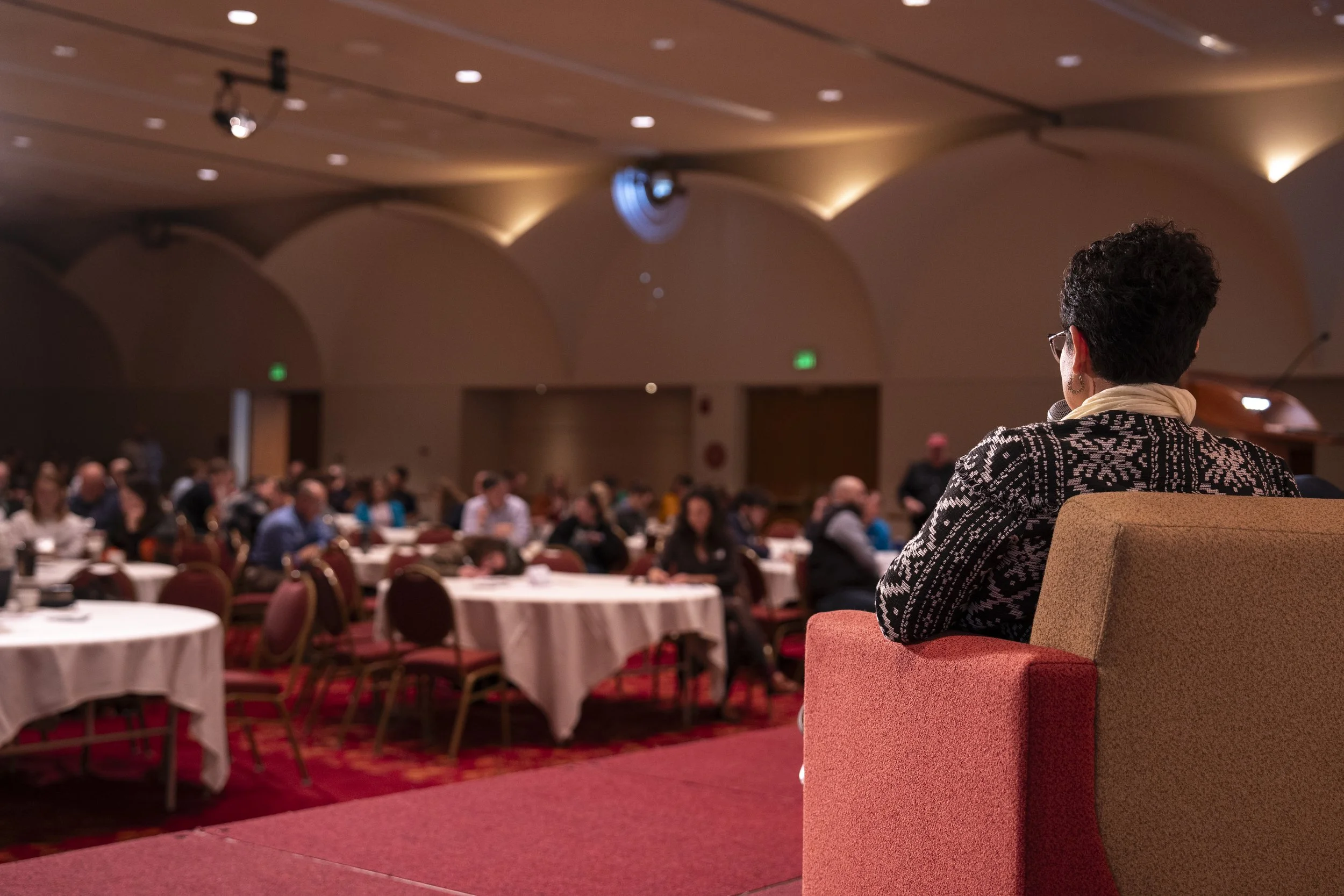 A woman with short curly hair, wearing glasses and a patterned jacket, seated on a stage or platform facing a large conference room filled with seated attendees at round tables.