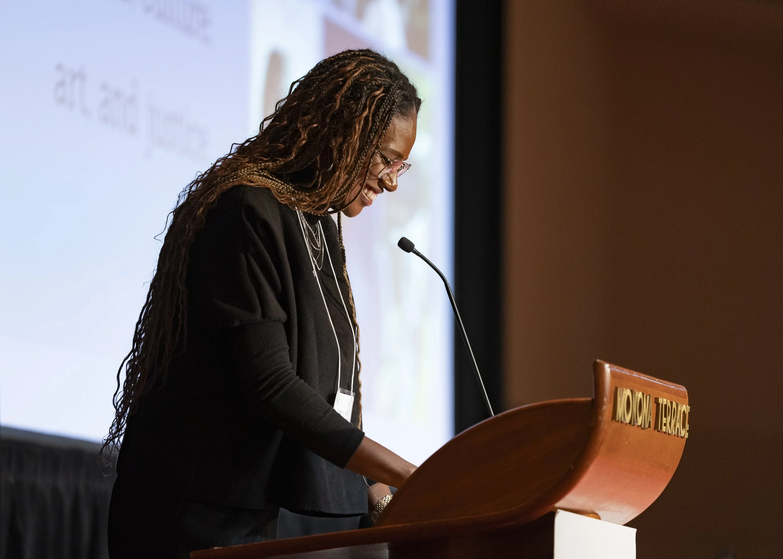 Woman with long braided hair and glasses speaking at a podium in a conference room