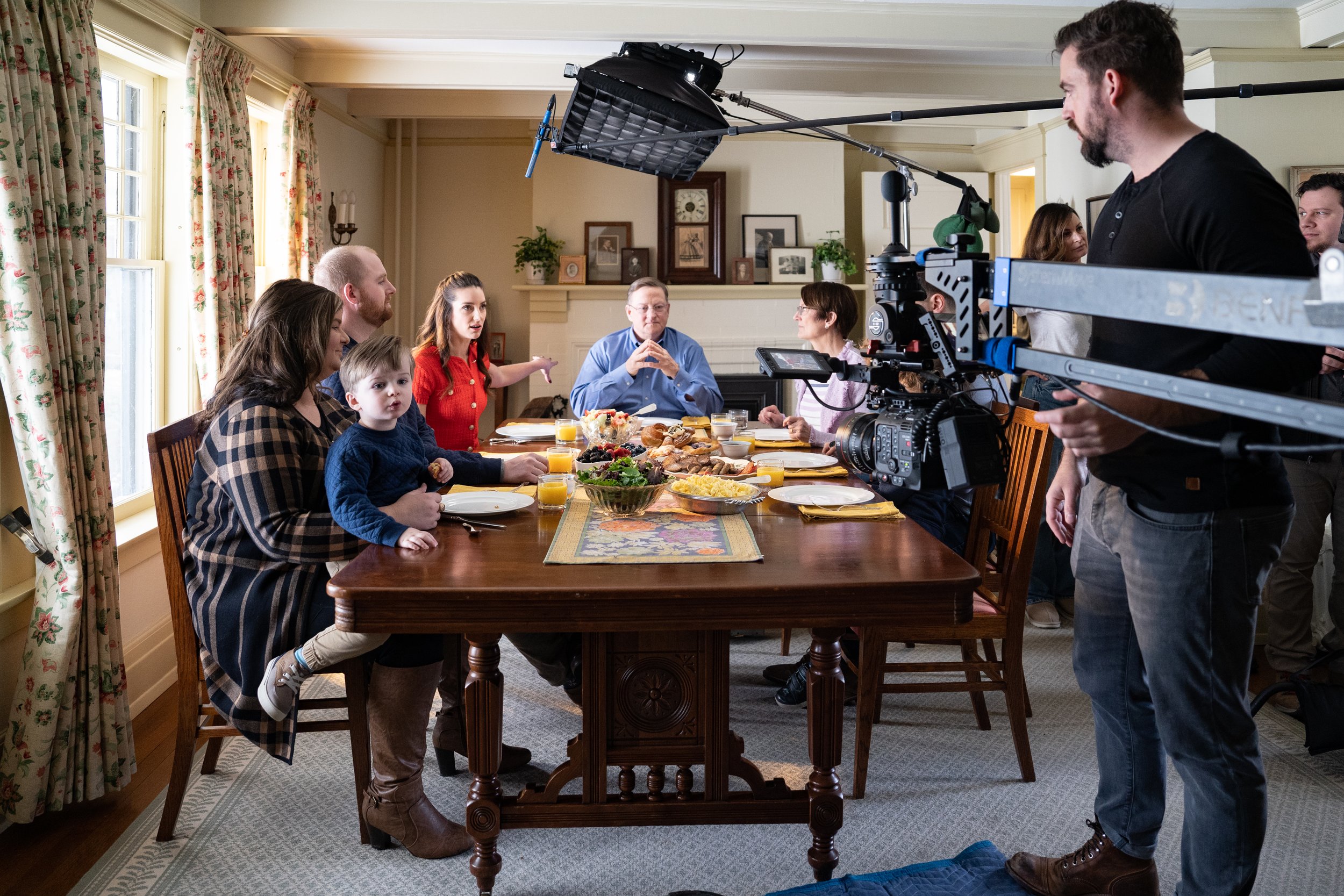 Filming a family breakfast scene in a well-lit dining room with a camera crew and a video camera pointed at a family of nine seated at a dining table with food and drinks.