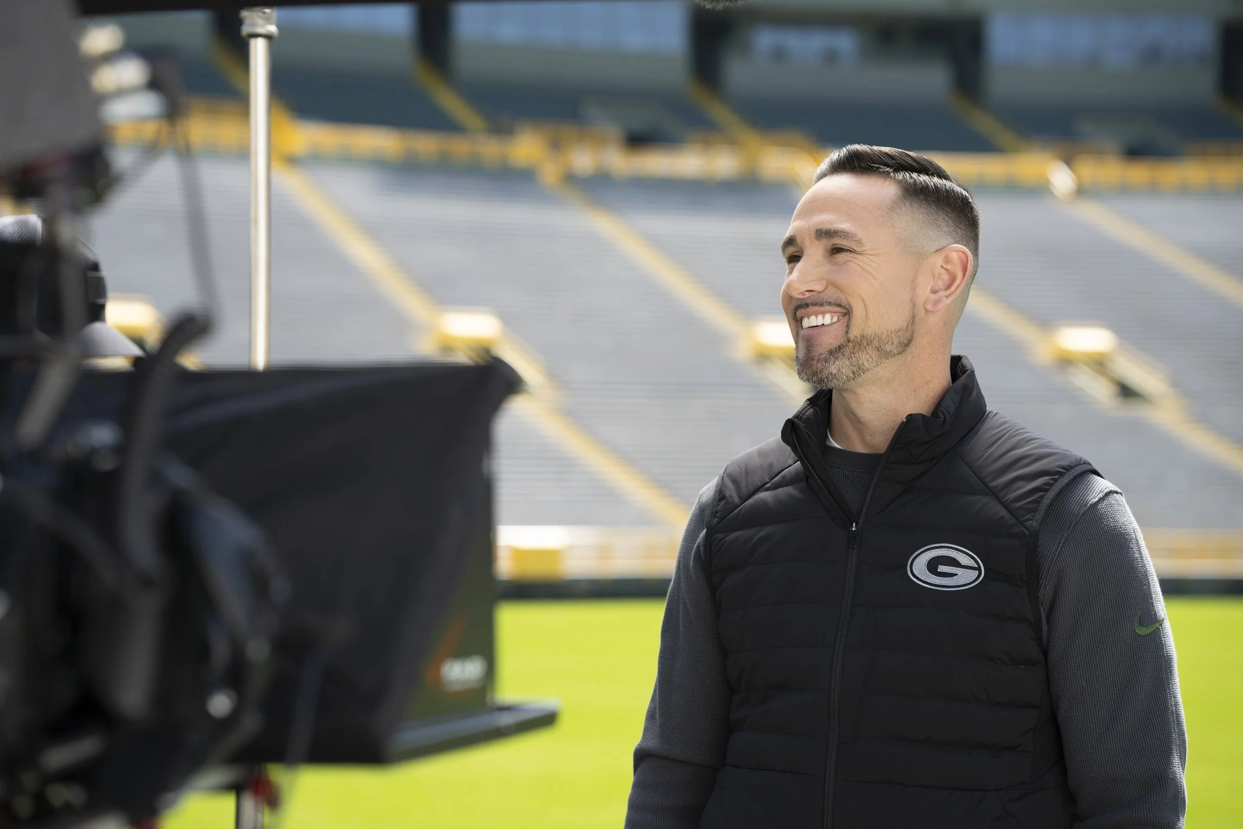 A man with short dark hair, beard, and mustache smiling and being interviewed at a sports stadium. He is wearing a black vest with an NFL Green Bay Packers logo and a gray long-sleeve shirt. The stadium seats are visible in the background.