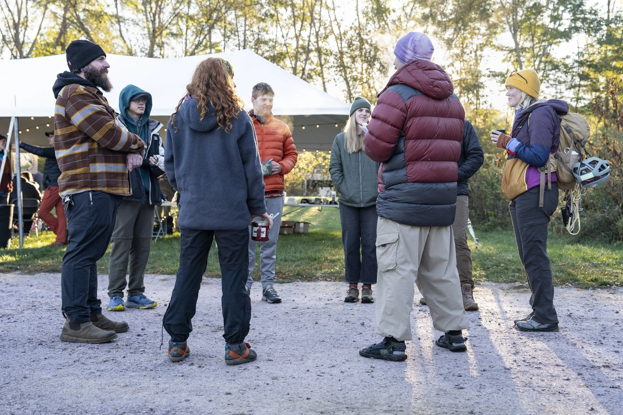 Group of people dressed in outdoor clothing standing in a circle outdoors near a white tent, engaging in conversation during daytime, surrounded by trees with sunlight filtering through.