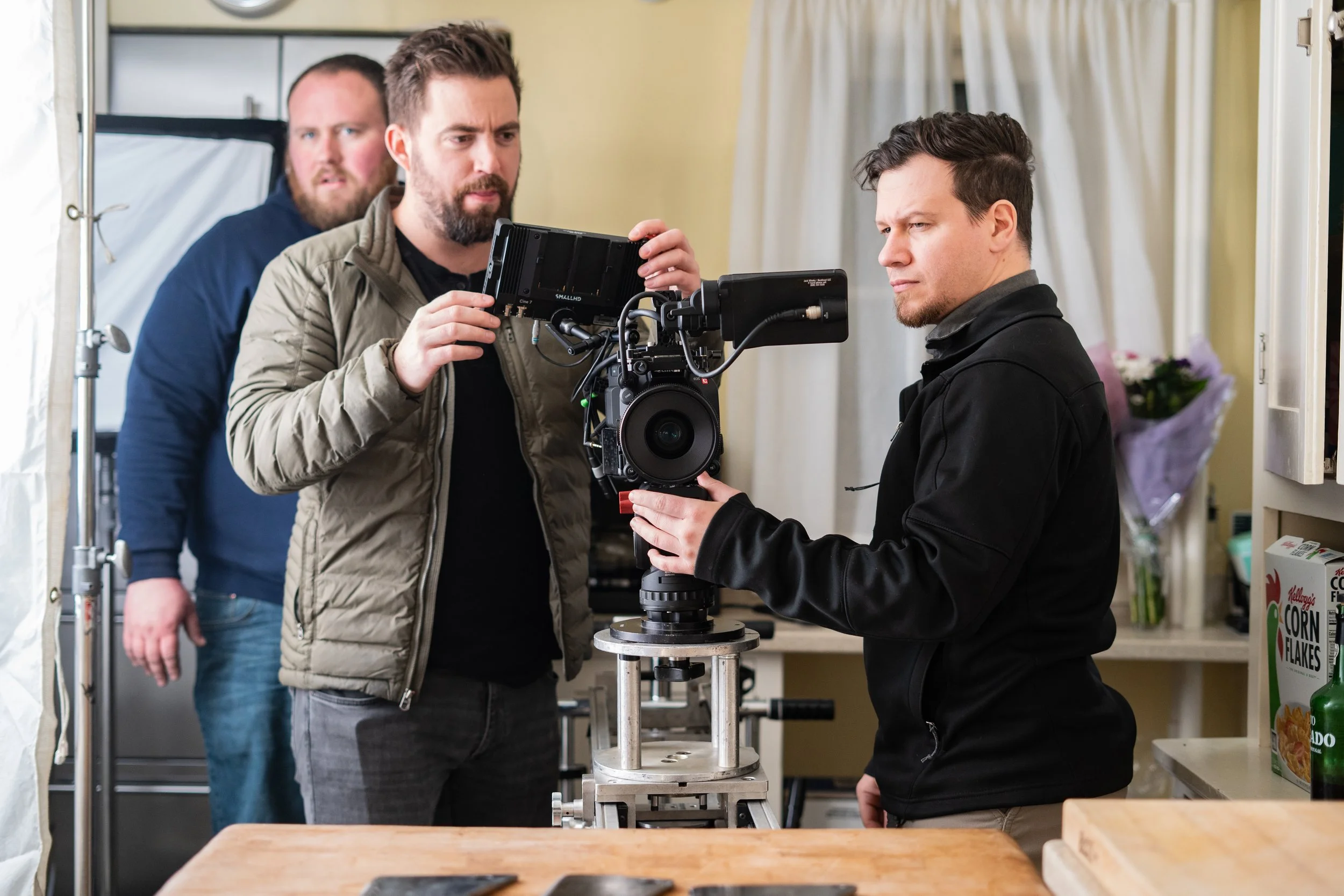 Three men working on camera setup in a kitchen, with one man adjusting the camera on a tripod, while the other two observe.
