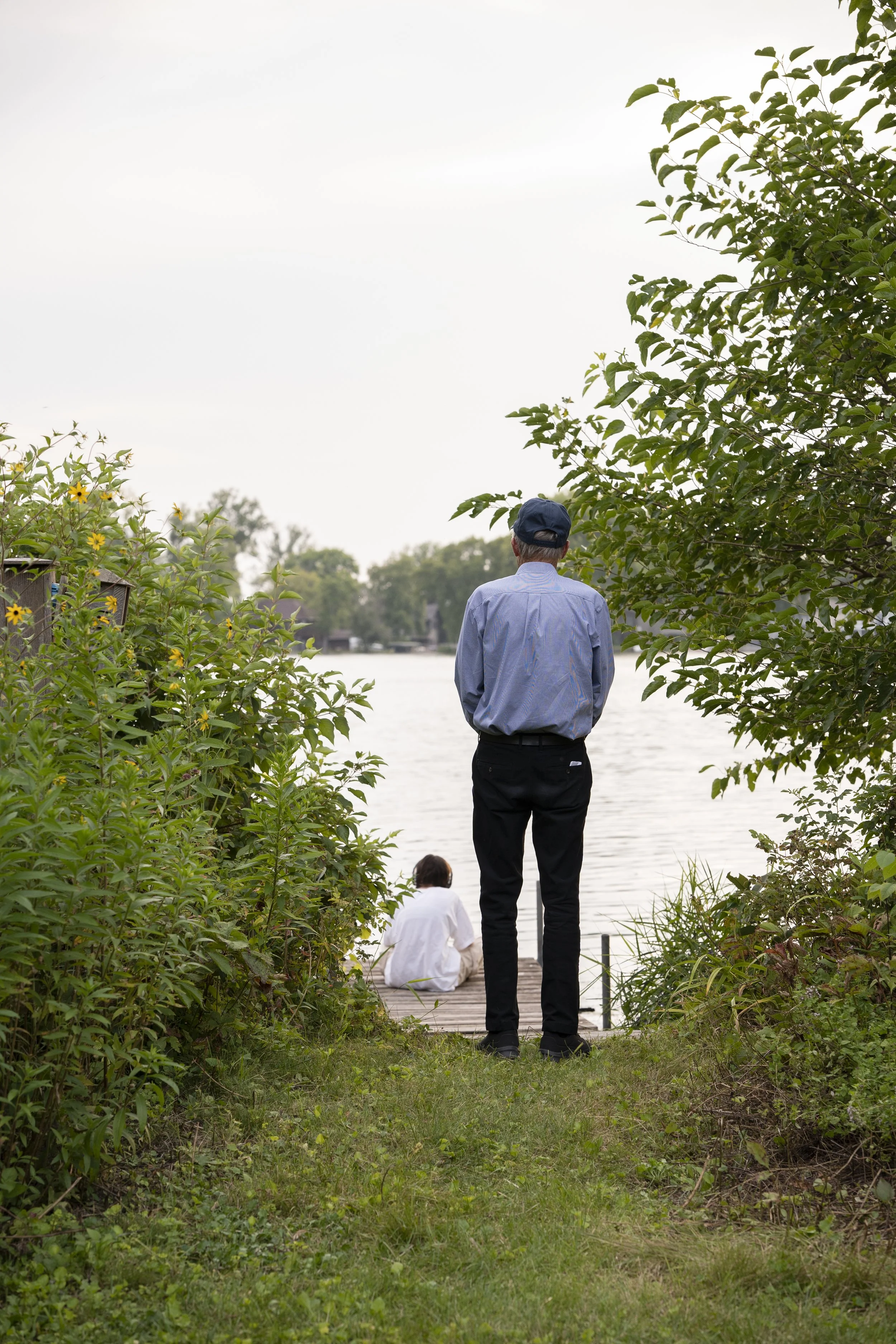 An older man and a woman by the water, with the man standing and the woman sitting on a dock, surrounded by greenery.